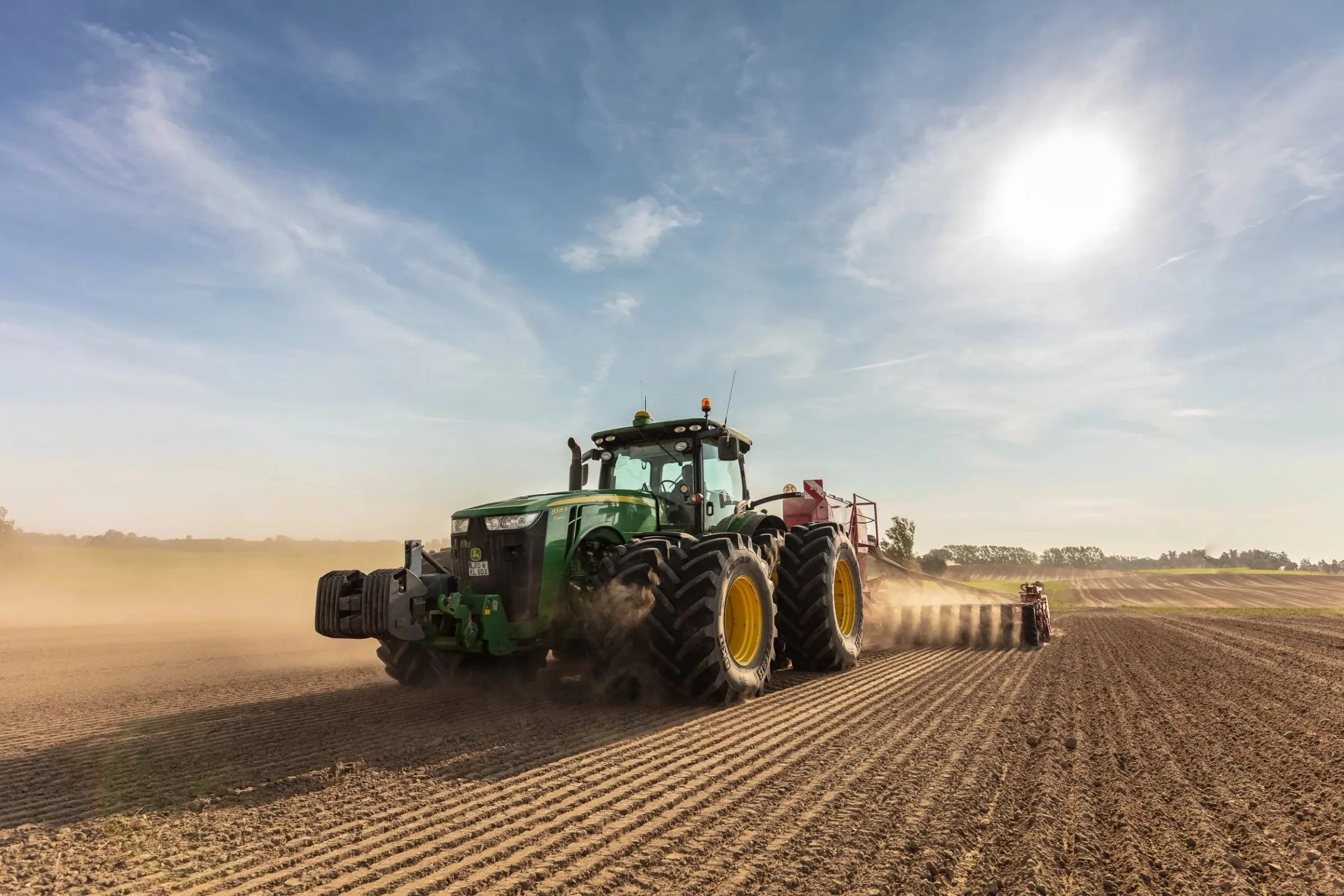 Grüner Traktor mit Sägerät zieht eine Staubfahne über ein offenes Feld im Gegenlicht