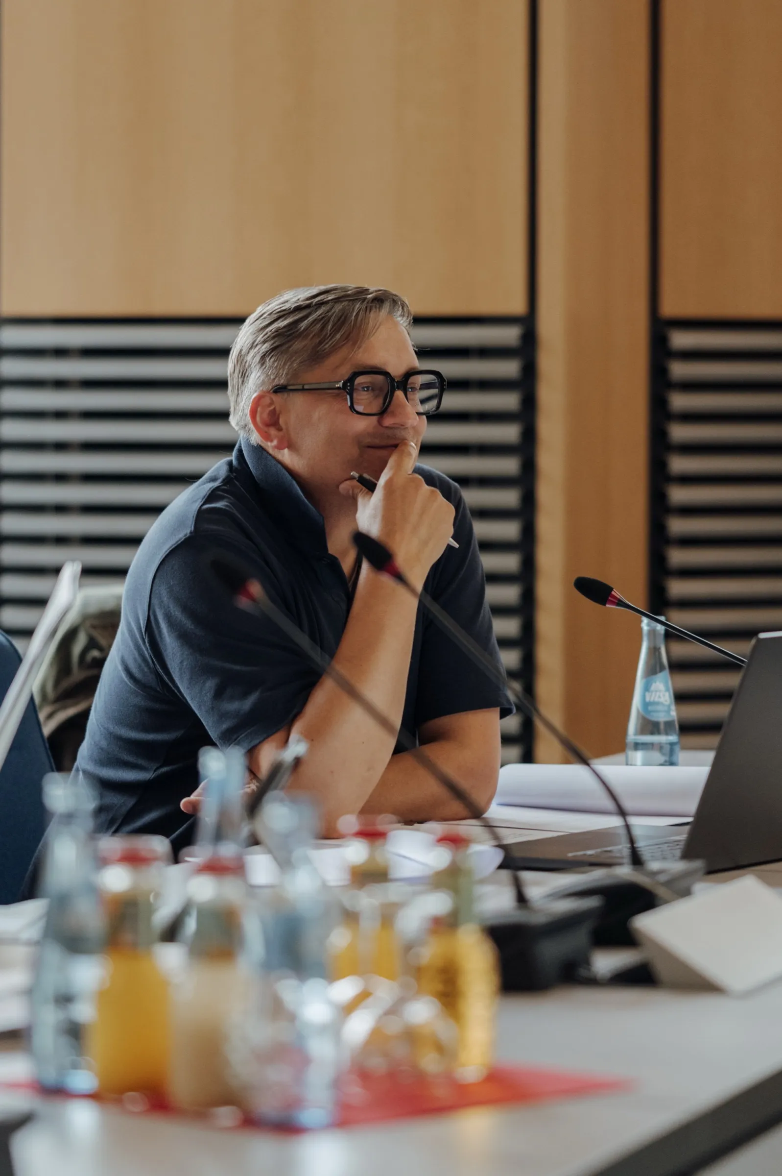 Roman Thomas lächelt am Sitzungstisch, die Hand am Kinn, im Vordergrund Wasser- und Saftflaschen