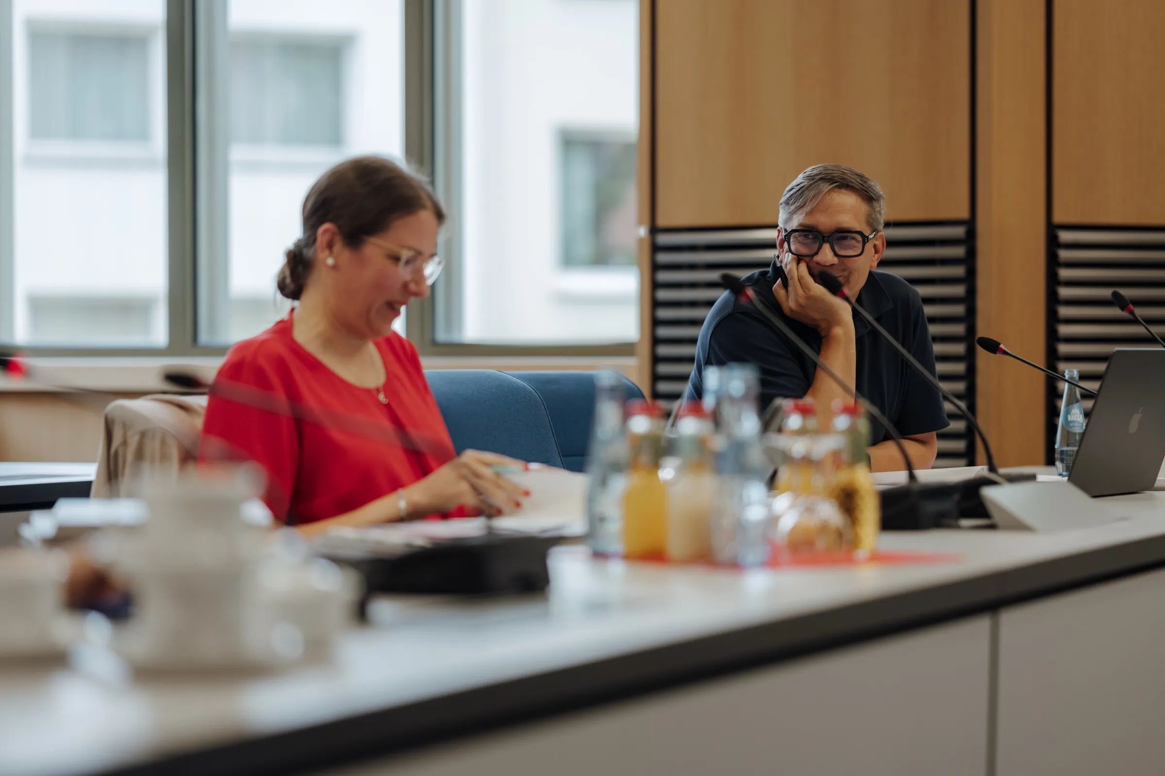 Dr. Anna Heinze und Roman Thomas am langen Sitzungstisch, vor ihnen Mikrofone, Wasser- und Saftflaschen, dahinter hohe Fenster und Holzwände