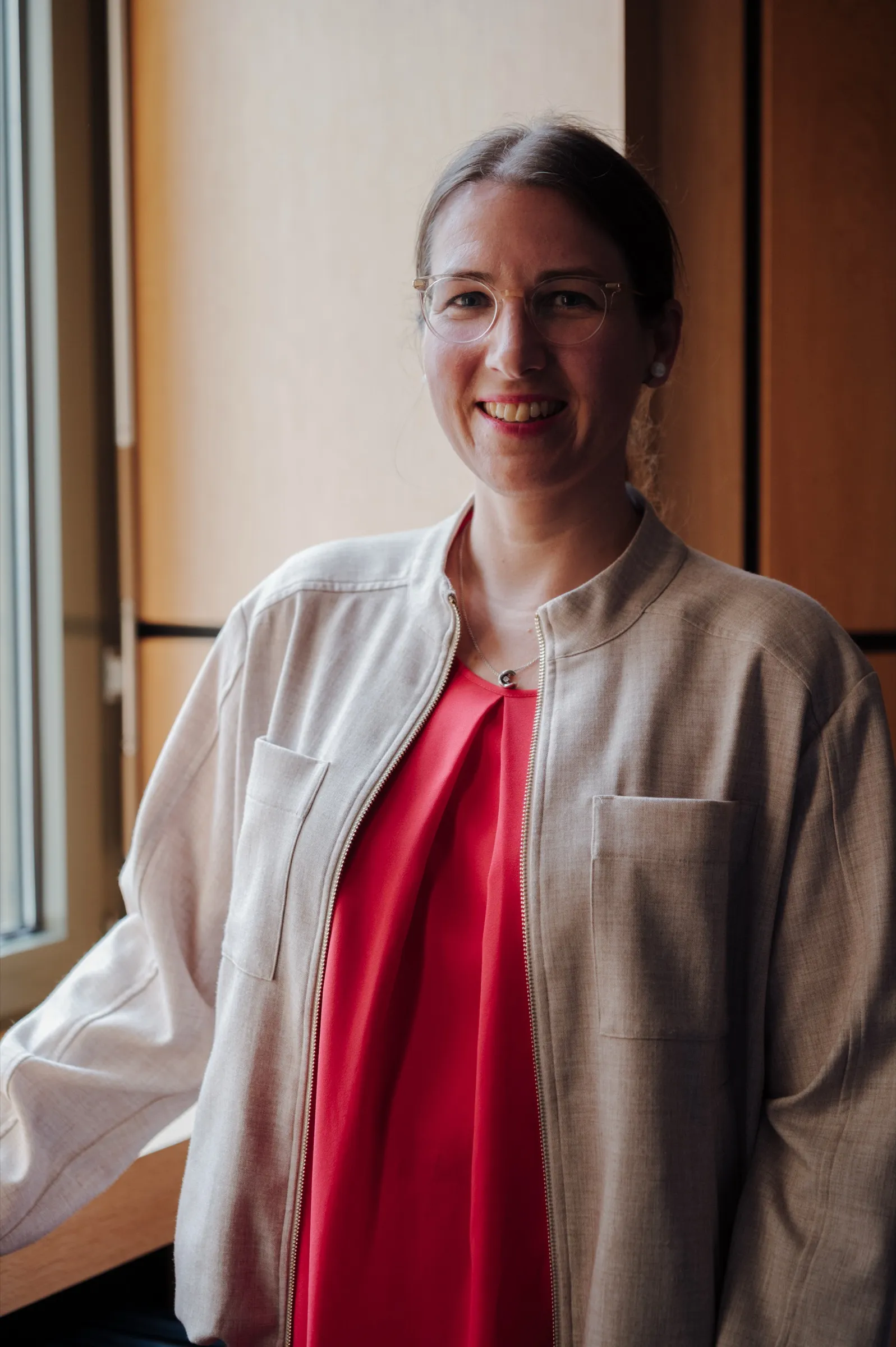 Porträt Dr. Anna Heinze am Fenster, rotes Oberteil und helle Jacke, sie lächelt in die Kamera