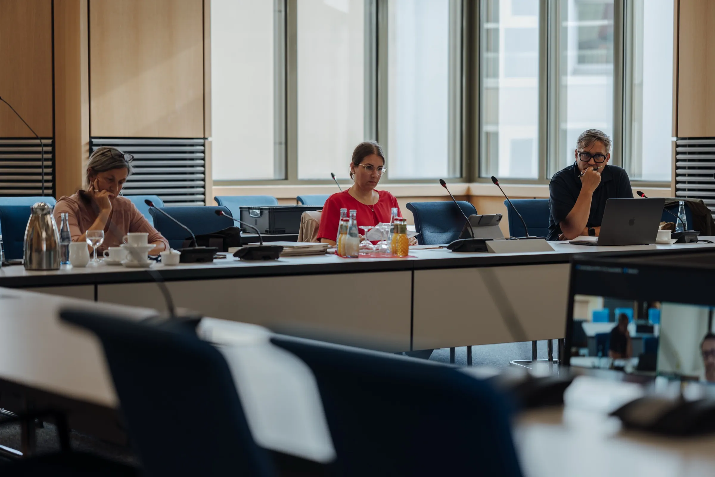 Der Sitzungssaal im Landtag: lange weiße Tische mit Mikrofonen und Flaschen, blaue Stühle, hohe Fenster, am Tisch drei Jurymitglieder