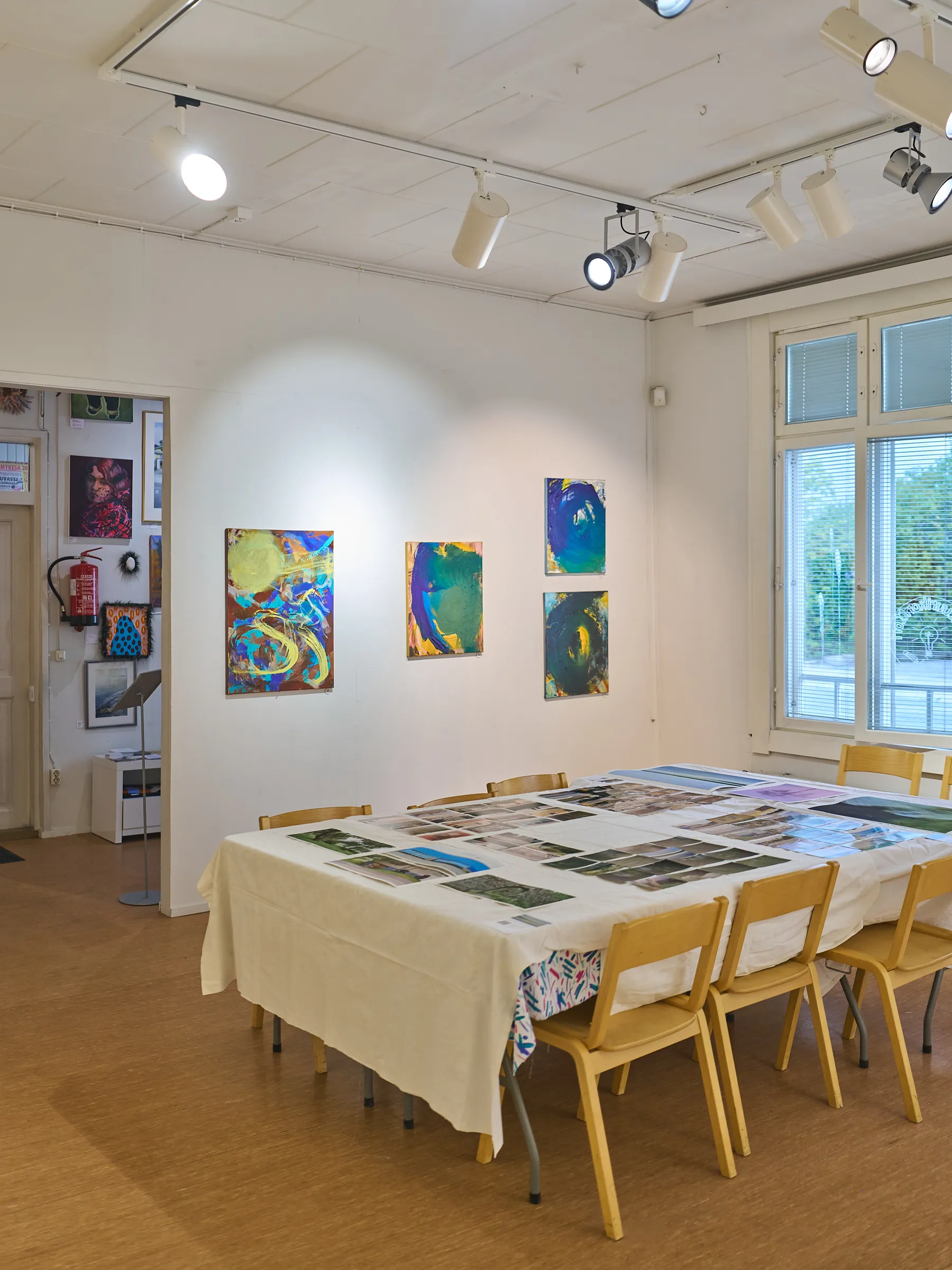 Heller Ausstellungsraum mit Schienenstrahlern, farbigen Gemälden an der Wand und einem langen Tisch mit ausgelegten Drucken
