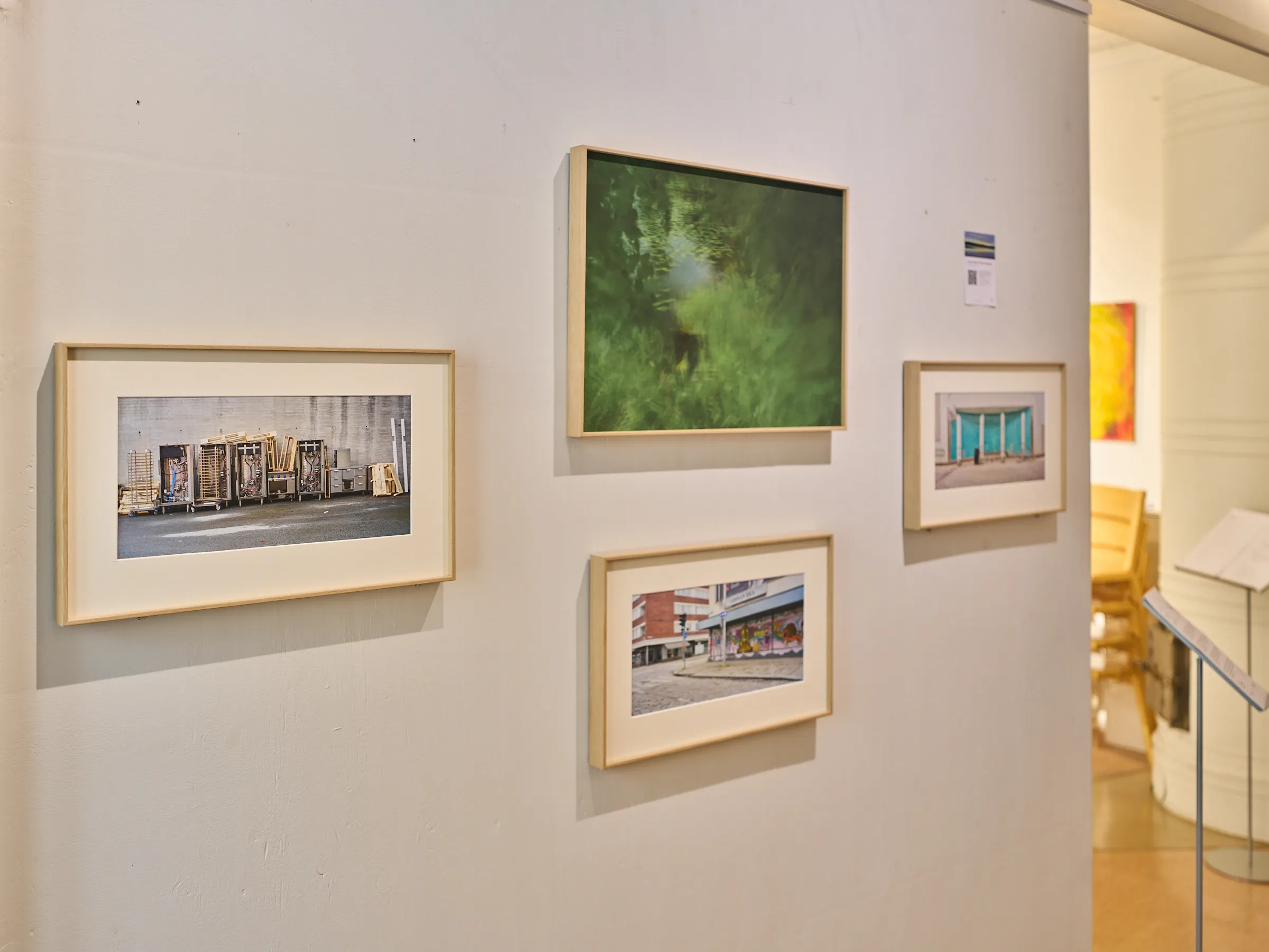 Blick an der Galeriewand entlang, gerahmte Fotografien und dahinter der helle Raum mit gelben Stühlen