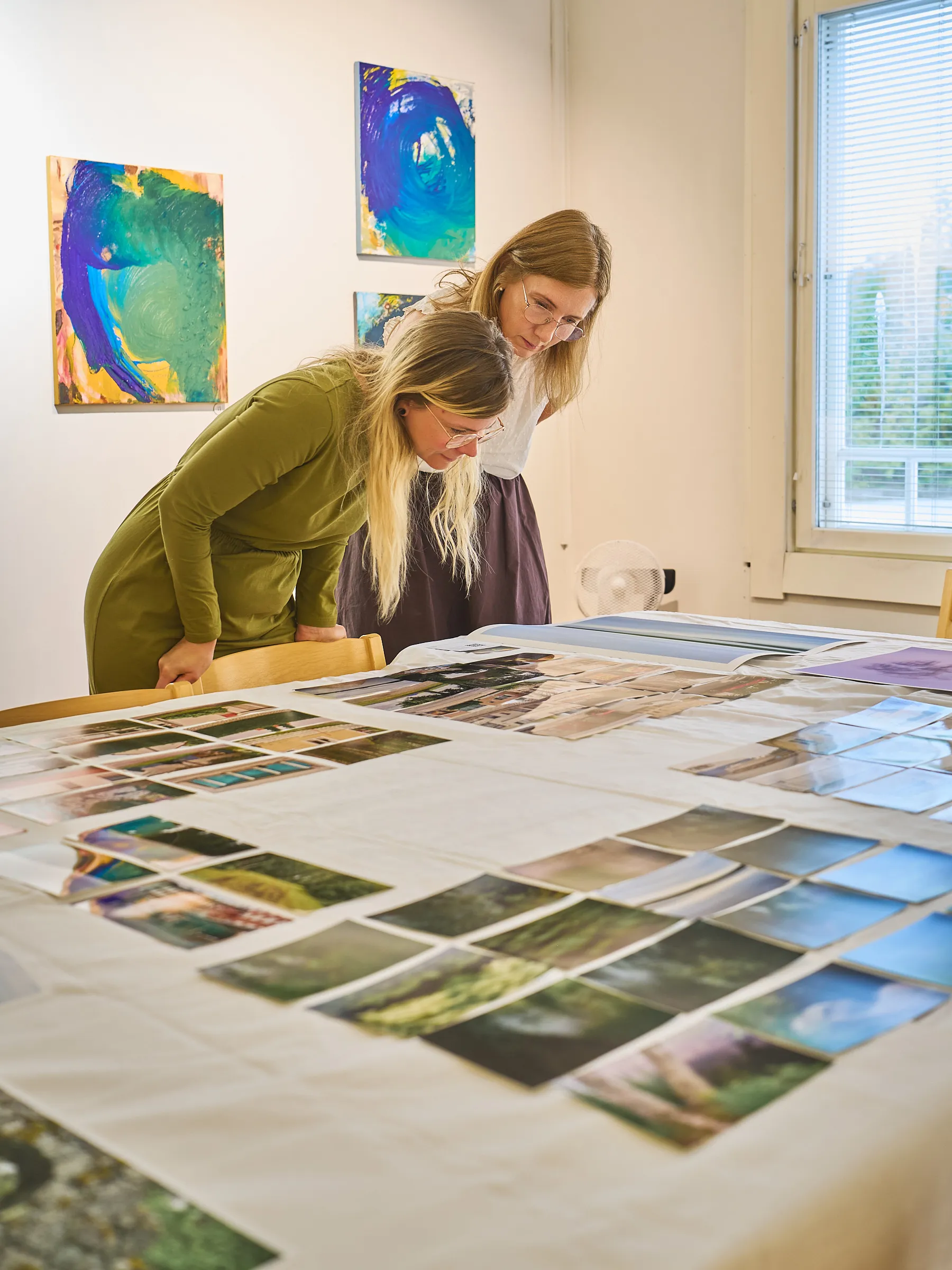 Zwei Frauen beugen sich dicht über die Bilder auf dem Tisch, im Hintergrund farbige Gemälde