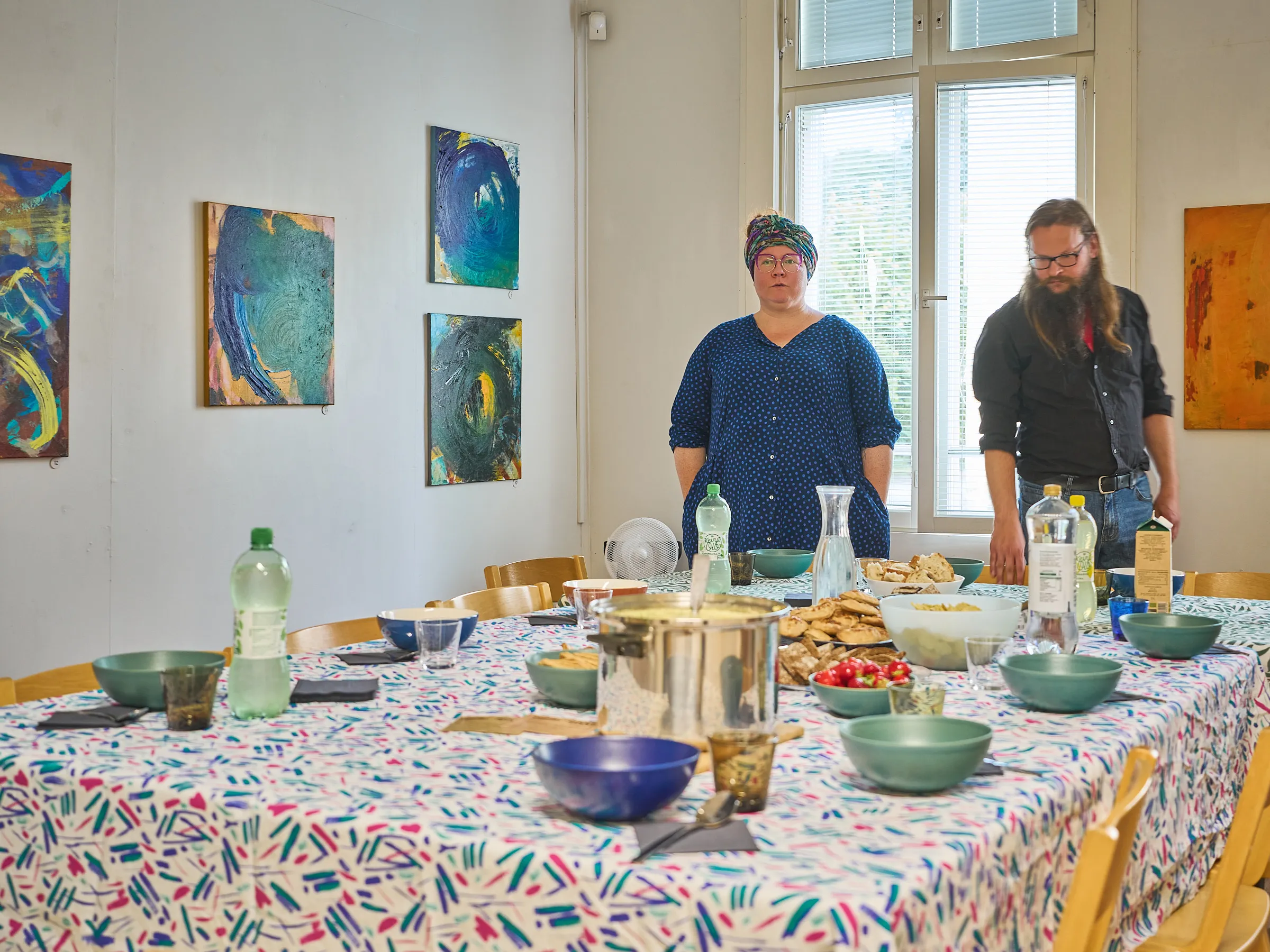 Gedeckter Tisch mit buntem Tuch, Schalen und Flaschen, dahinter stehen zwei Menschen im hellen Raum