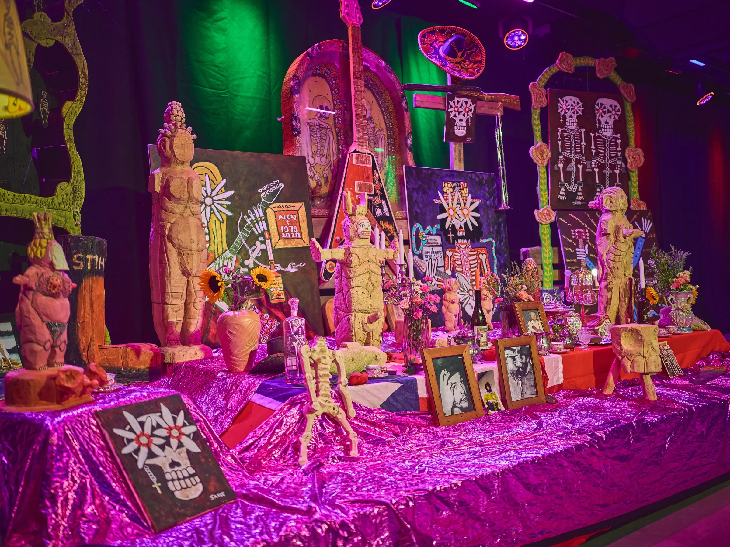 Reich geschmückter Erinnerungsaltar mit Figuren, Kerzen und gerahmten Fotos auf pink glänzendem Tuch