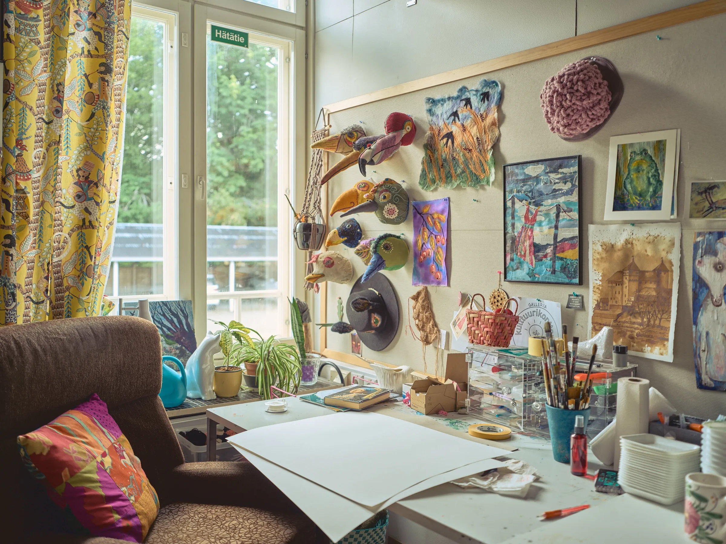 Atelier im Kulttuurikonttori: Stoffvogel-Masken, Filzarbeiten und Bilder an einer Pinnwand, davor ein Arbeitstisch mit Pinseln