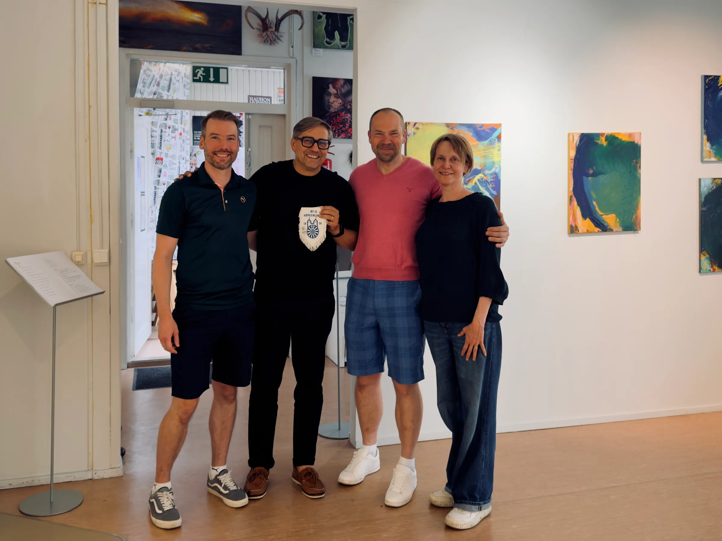 Vier Menschen stehen Arm in Arm in der kleinen Ausstellung, einer hält den Wimpel des RT-8 Hämeenlinna