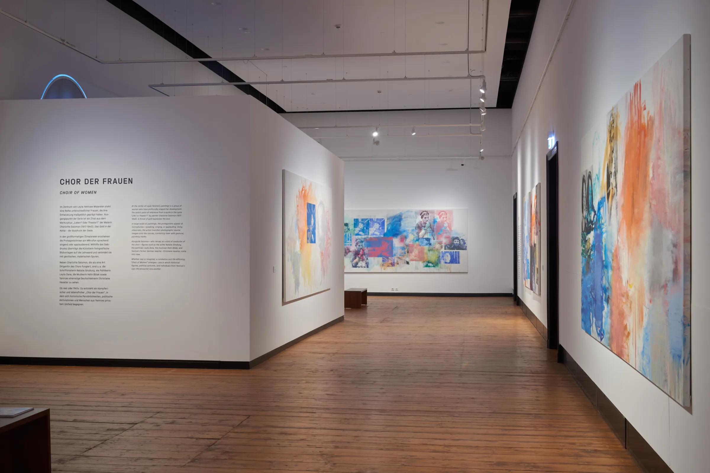 Ausstellungsraum mit Holzboden, links eine Wandtafel mit der Überschrift Chor der Frauen, an den Wänden große Gemälde in Blau und Orange