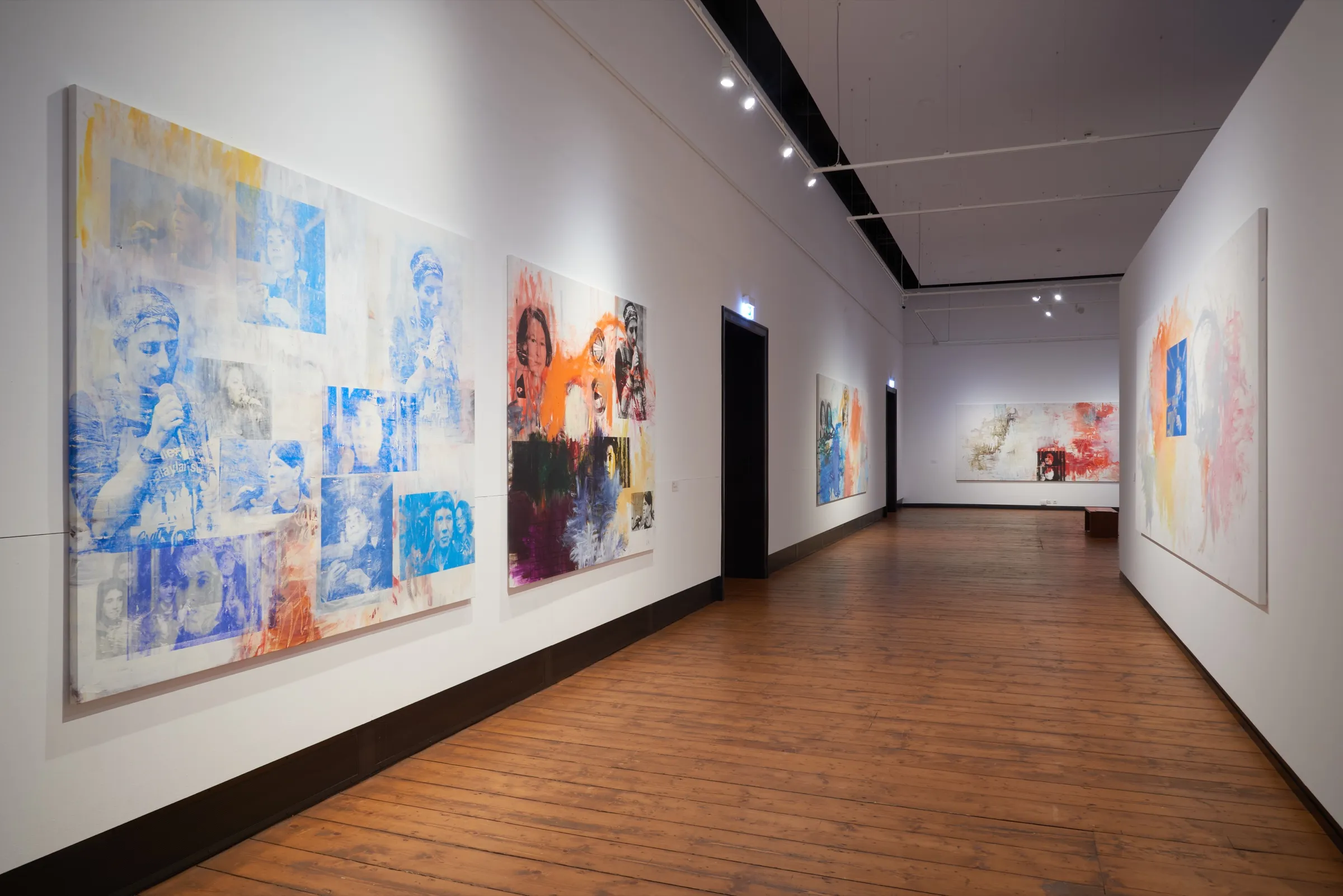 Langer Ausstellungsgang mit Holzdielen, an der linken Wand großformatige Gemälde mit blauen und orangen Siebdruckporträts