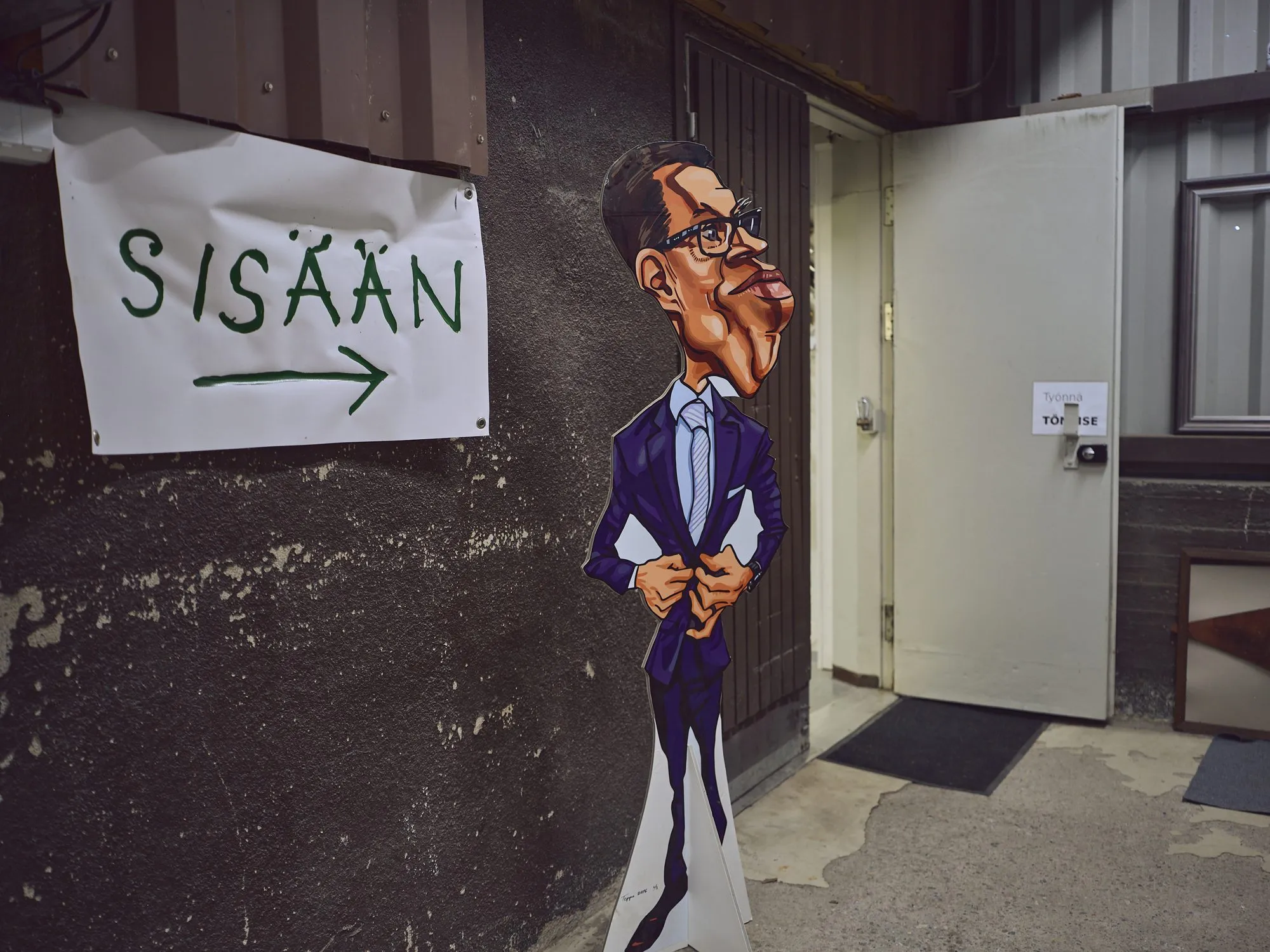 Drei sehr unterschiedliche Karikaturen von Alexander Stubb nebeneinander an einer Wand, als Spielkarte, als Ölbild und als Comicpaar