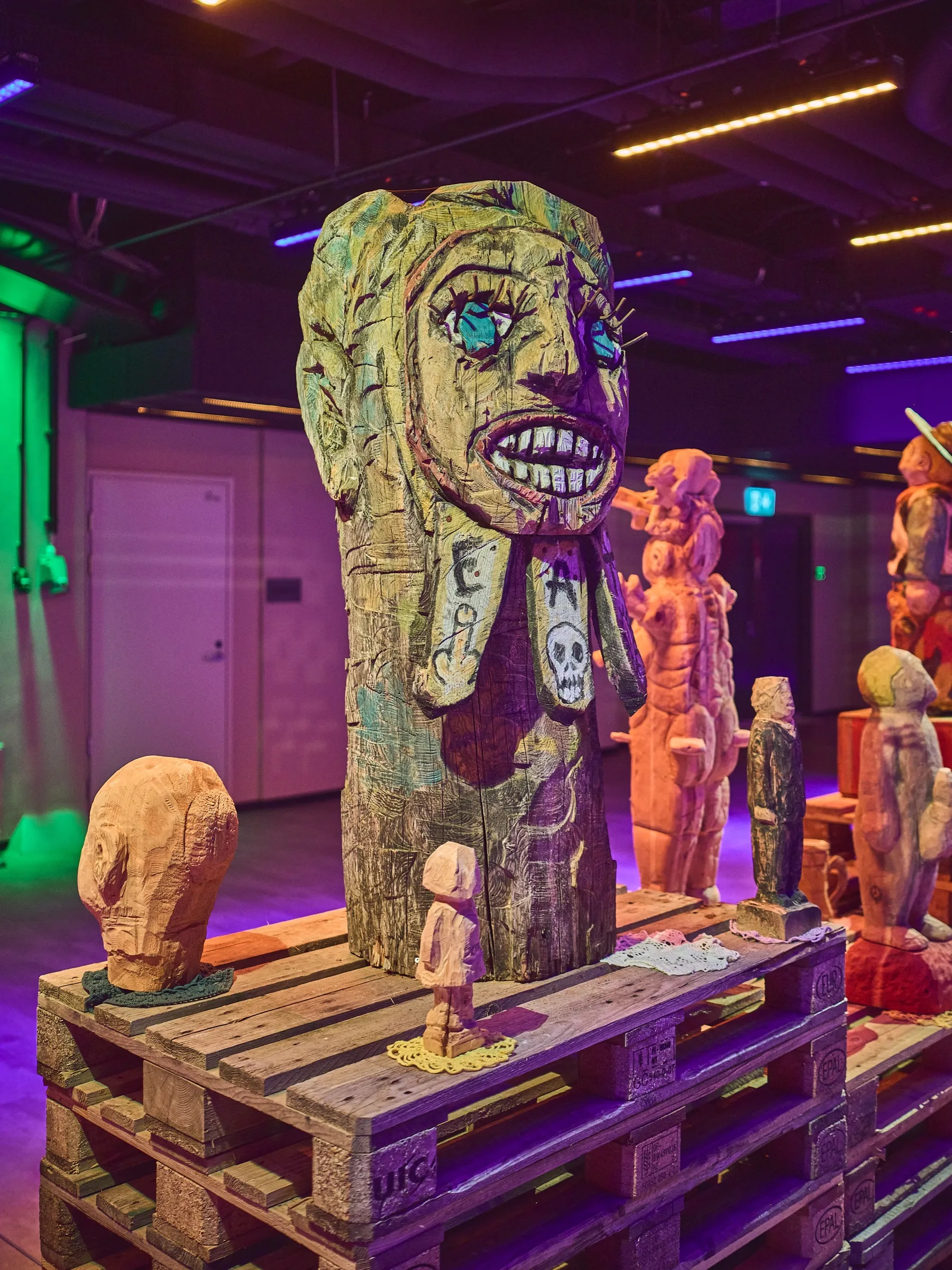 Nahe Holzfigur mit breitem Zähnegrinsen, grünlich beleuchtet, weitere Figuren dahinter