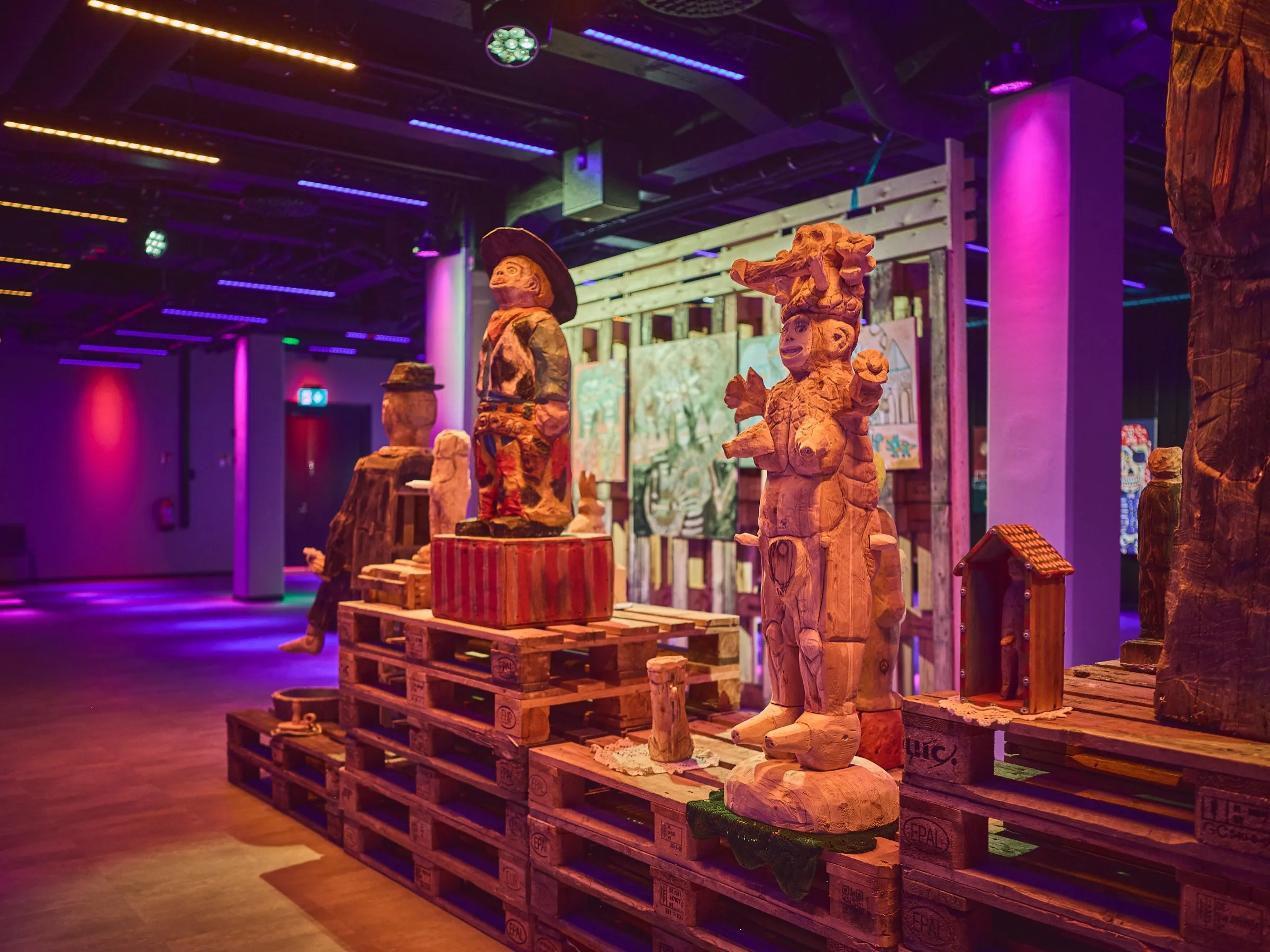 Gruppe hoher Holzfiguren auf Paletten zwischen violett beleuchteten Säulen