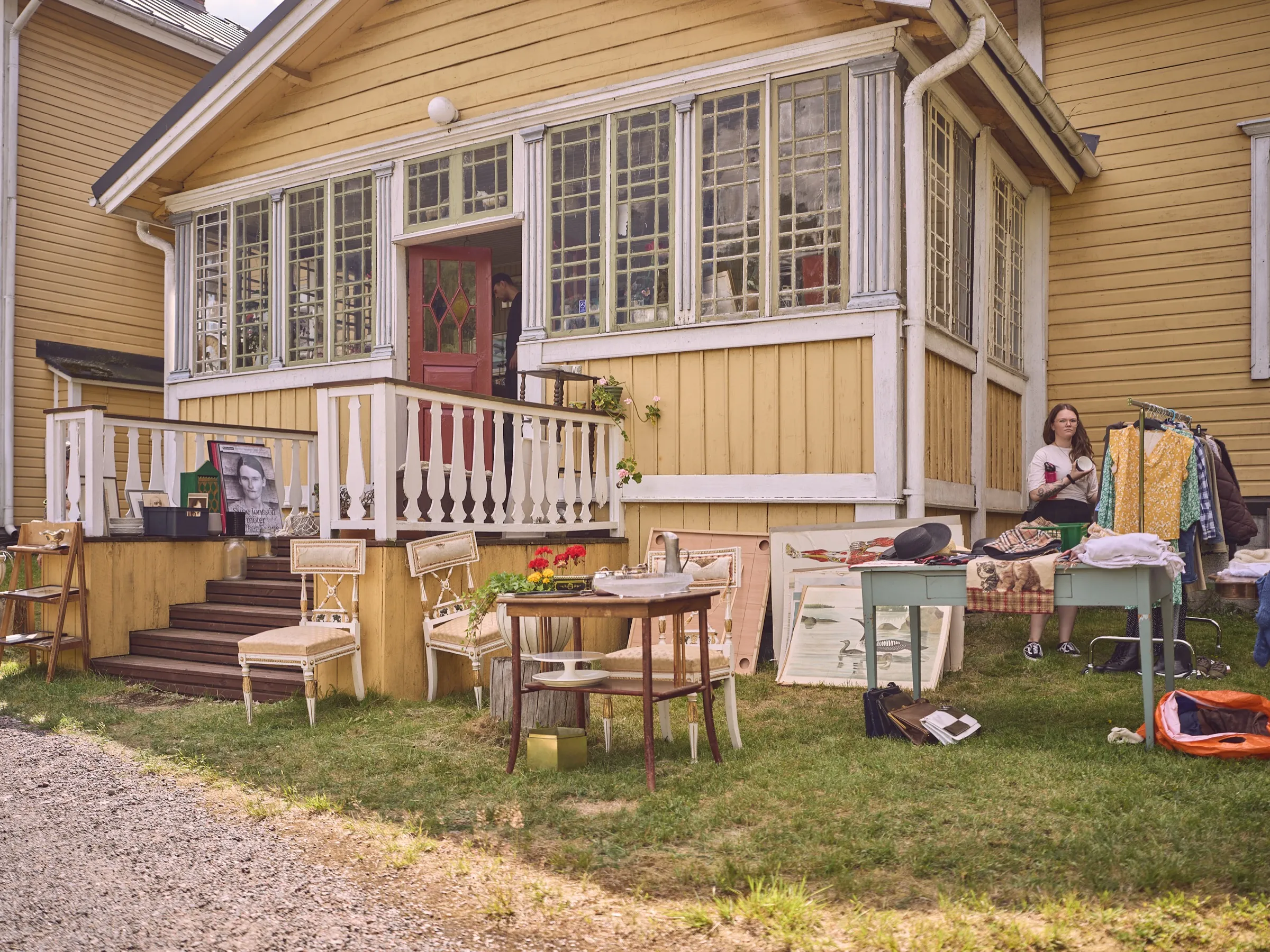 Gelbes Holzhaus mit Veranda und Treppe, davor Flohmarktmöbel im Gras