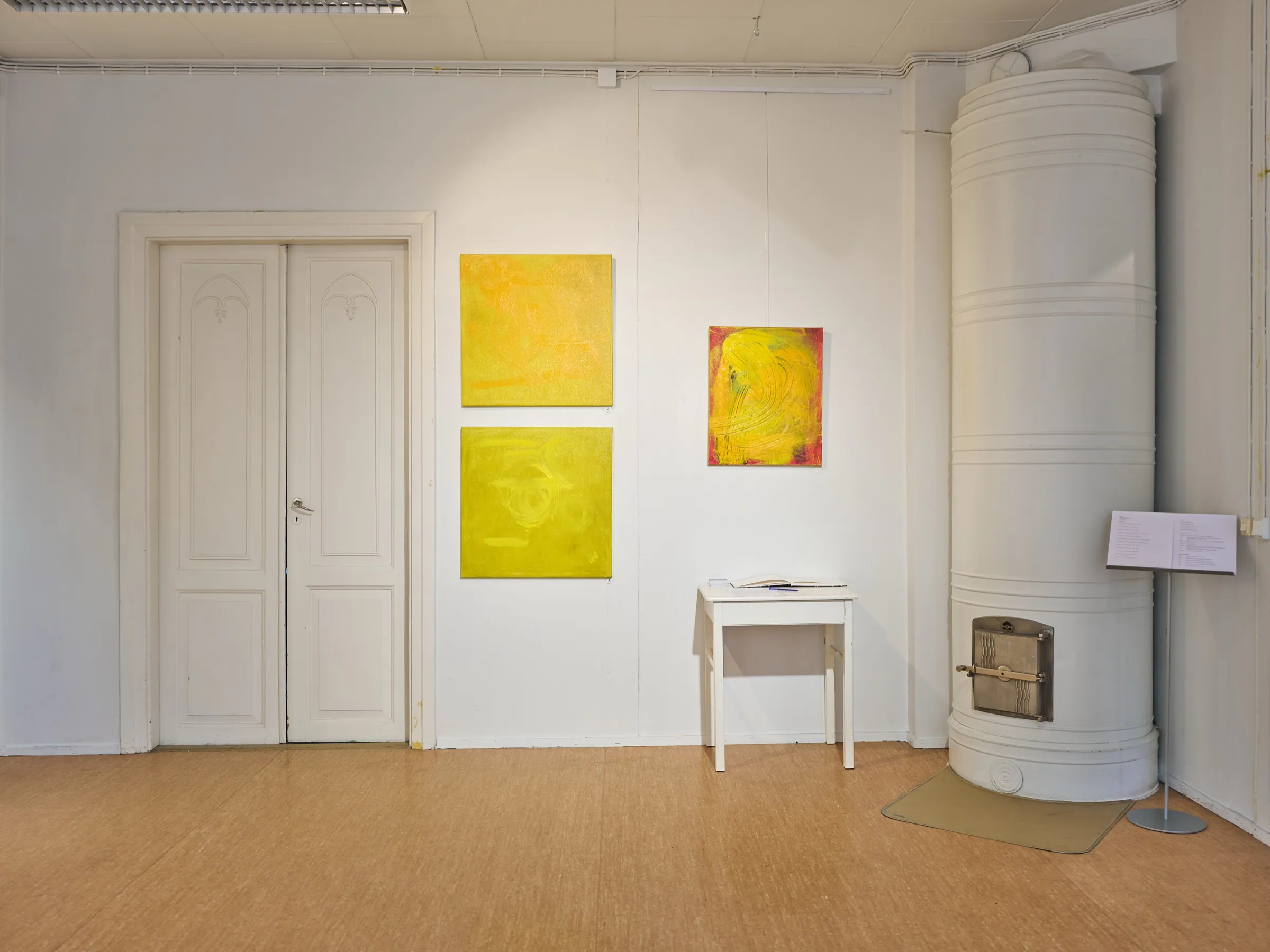 Galerieraum im Kulttuurikonttori mit gelben abstrakten Bildern an weißer Wand und einem alten Kachelofen