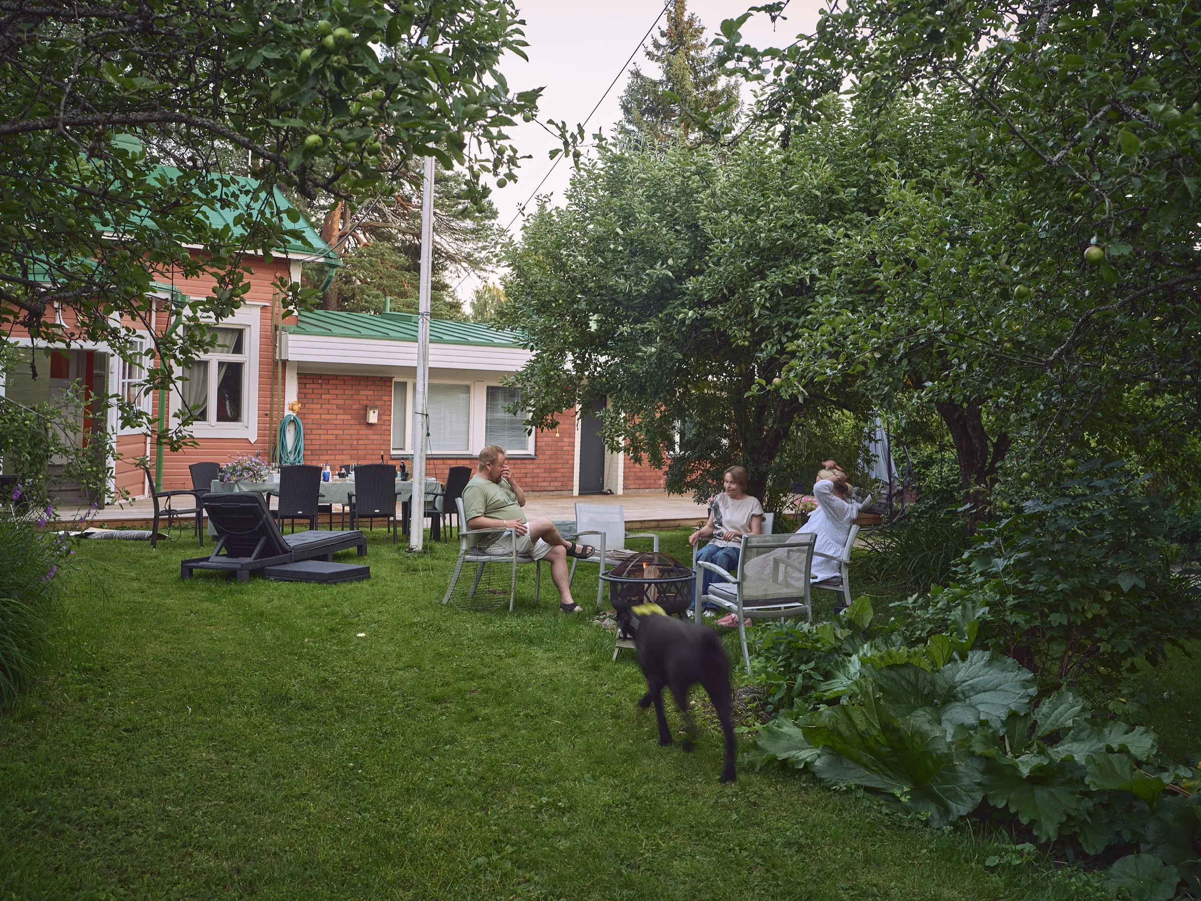 Abendliche Gartenrunde mit Stühlen, Menschen und einem Hund vor einem Backsteinhaus