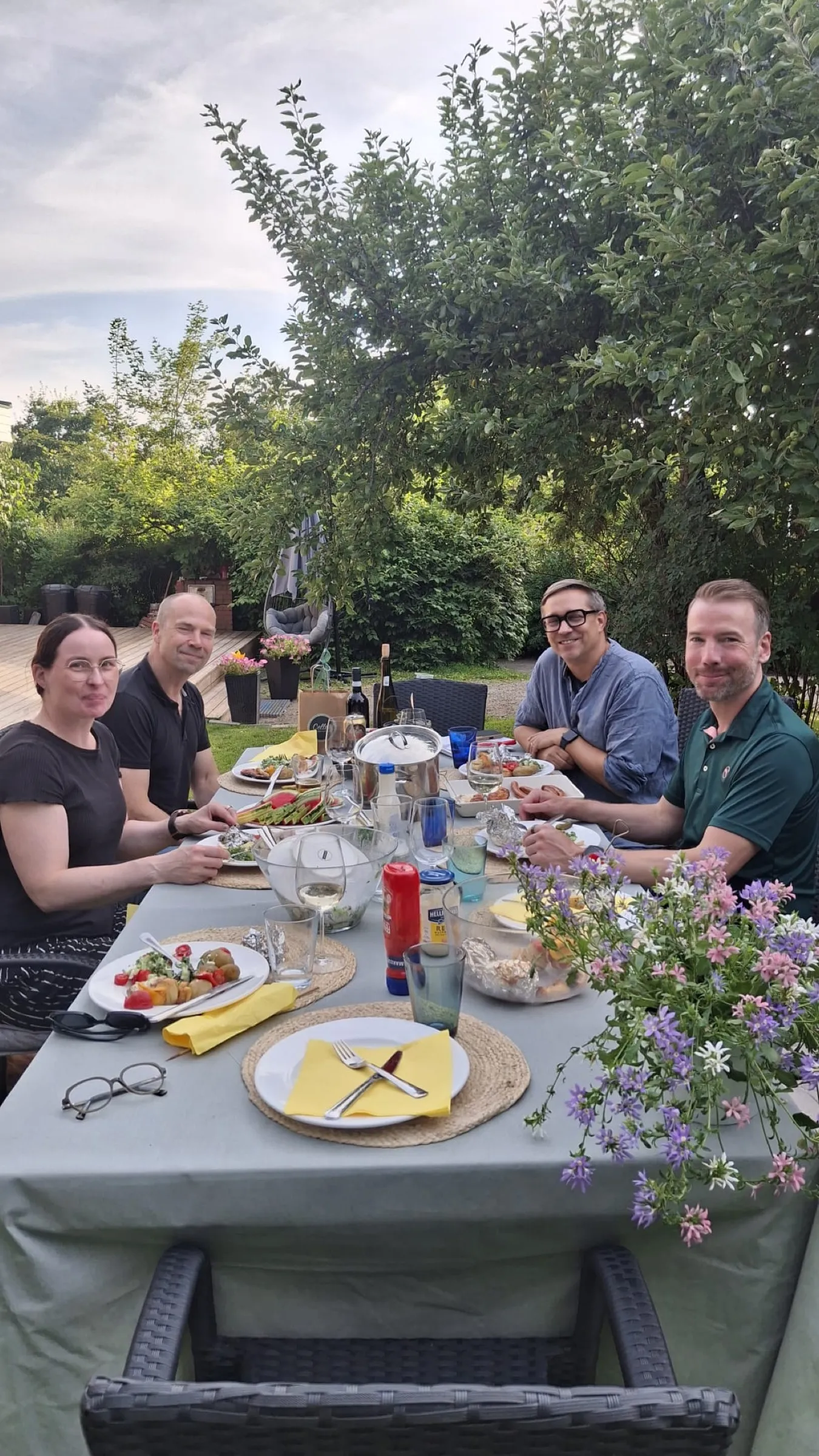 Sommerliche Tafel im Garten, vier Menschen beim Essen, Blumen im Vordergrund, Hochformat