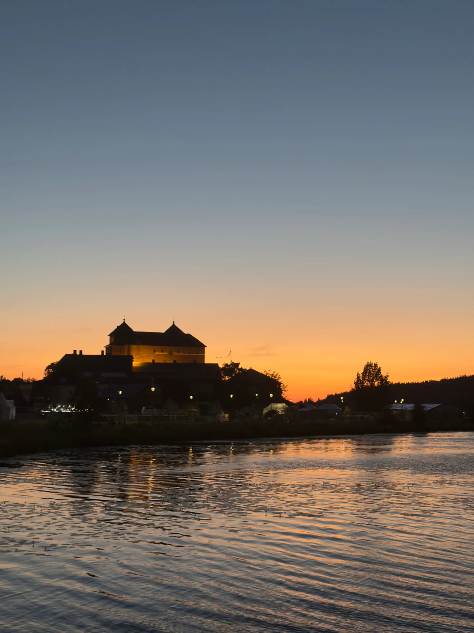 Die Burg von Hämeenlinna als dunkle Silhouette über dem See, dahinter ein orange glühender Nachthimmel, Hochformat