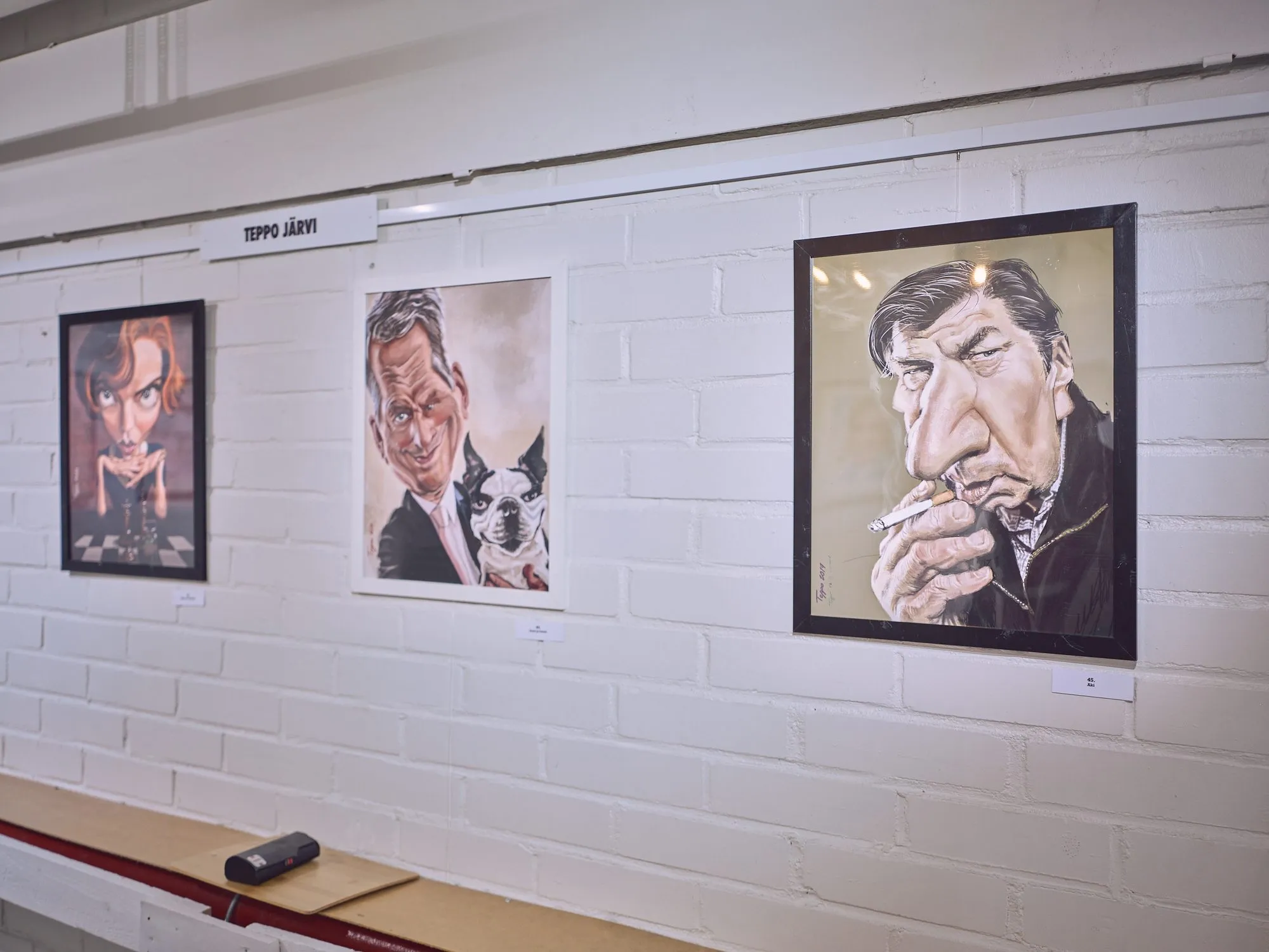 Drei Karikaturen von Teppo Järvi an einer weißen Backsteinwand, in der Mitte Niinistö mit einem kleinen Hund