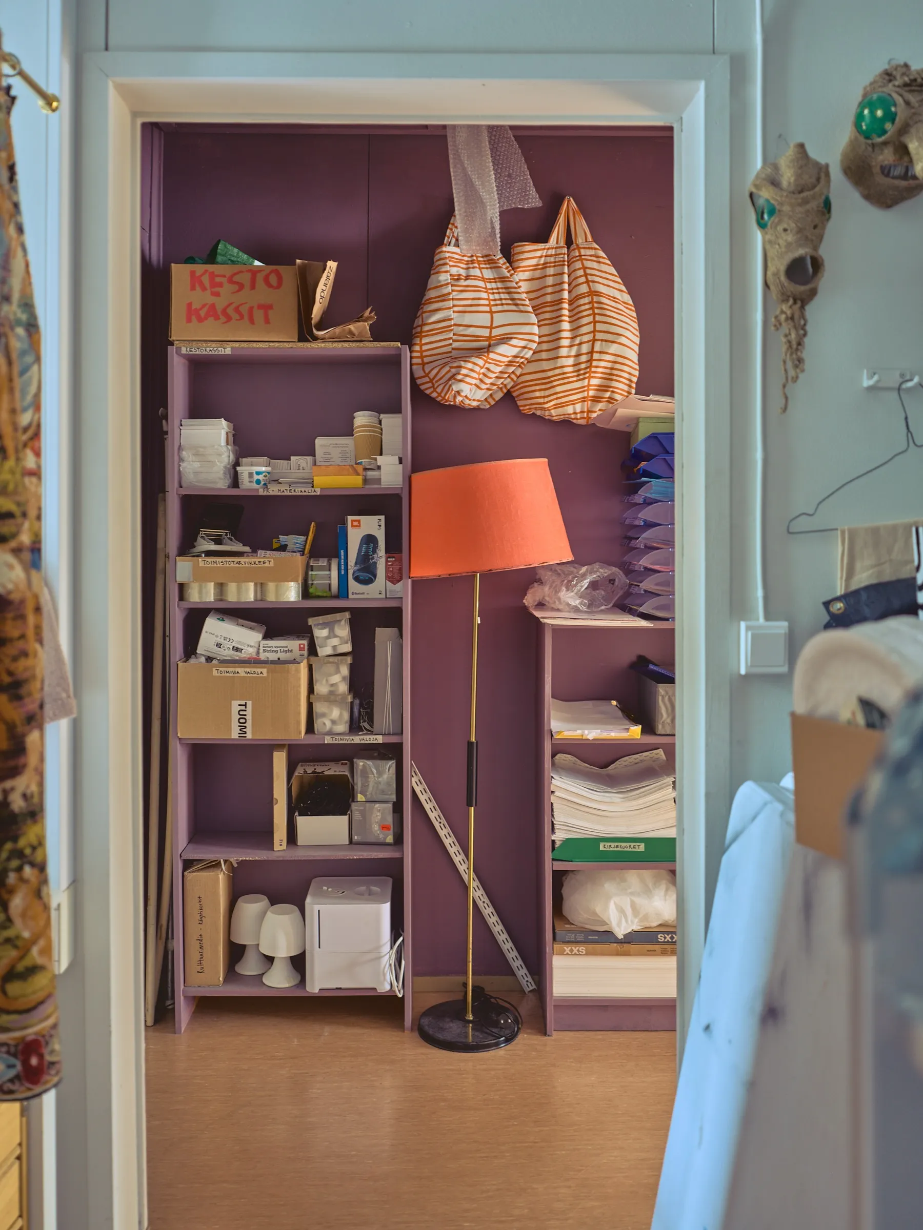 Blick ins Lager mit lila Regalen, Stofftaschen und einer Stehlampe mit orangefarbenem Schirm