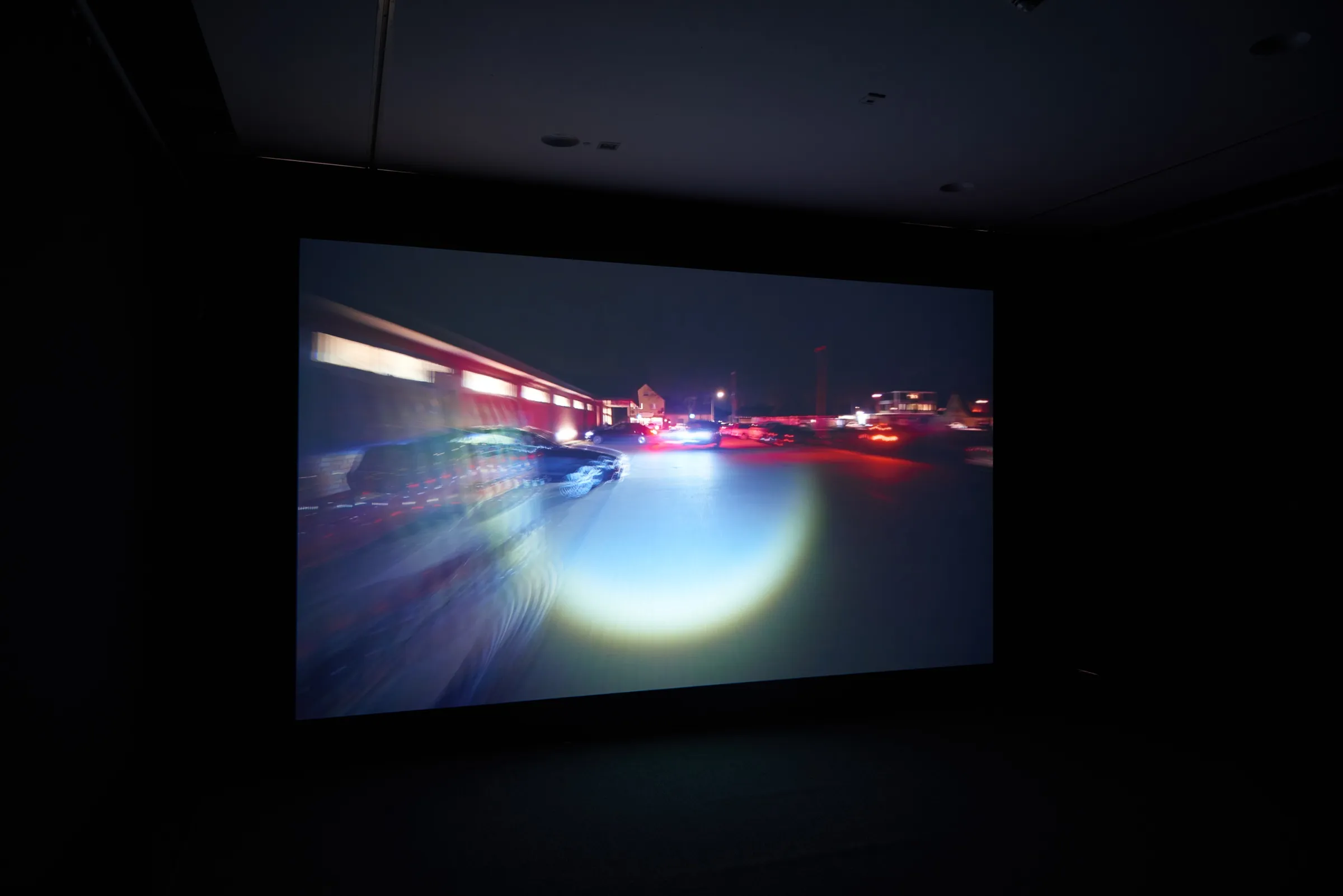 Dunkler Raum mit großer Projektion: eine nächtliche Straße, verwischte Lichter und ein heller Lichtkegel auf dem Asphalt