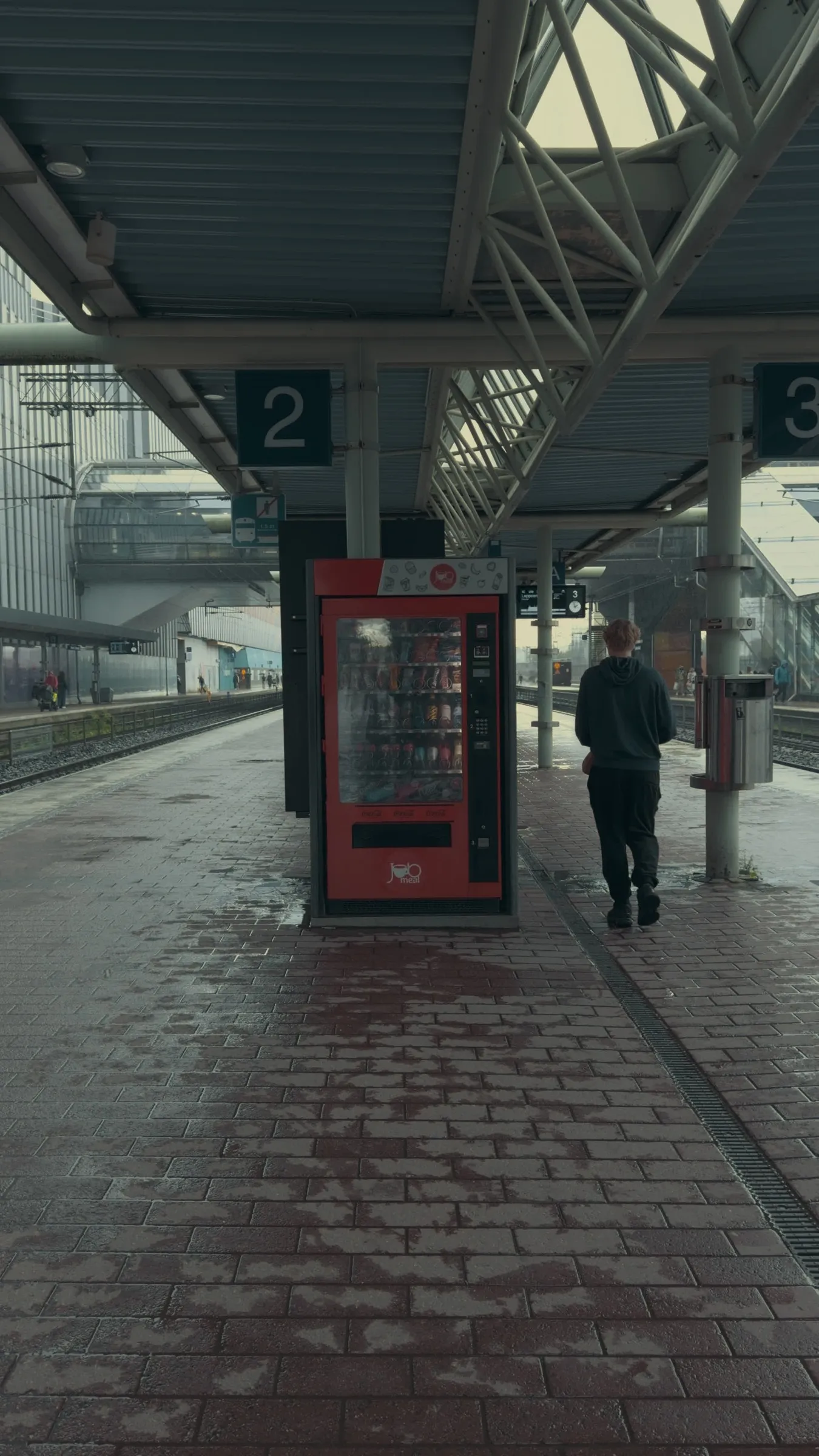 Verregneter Bahnsteig mit rotem Snackautomat, eine Person wartet