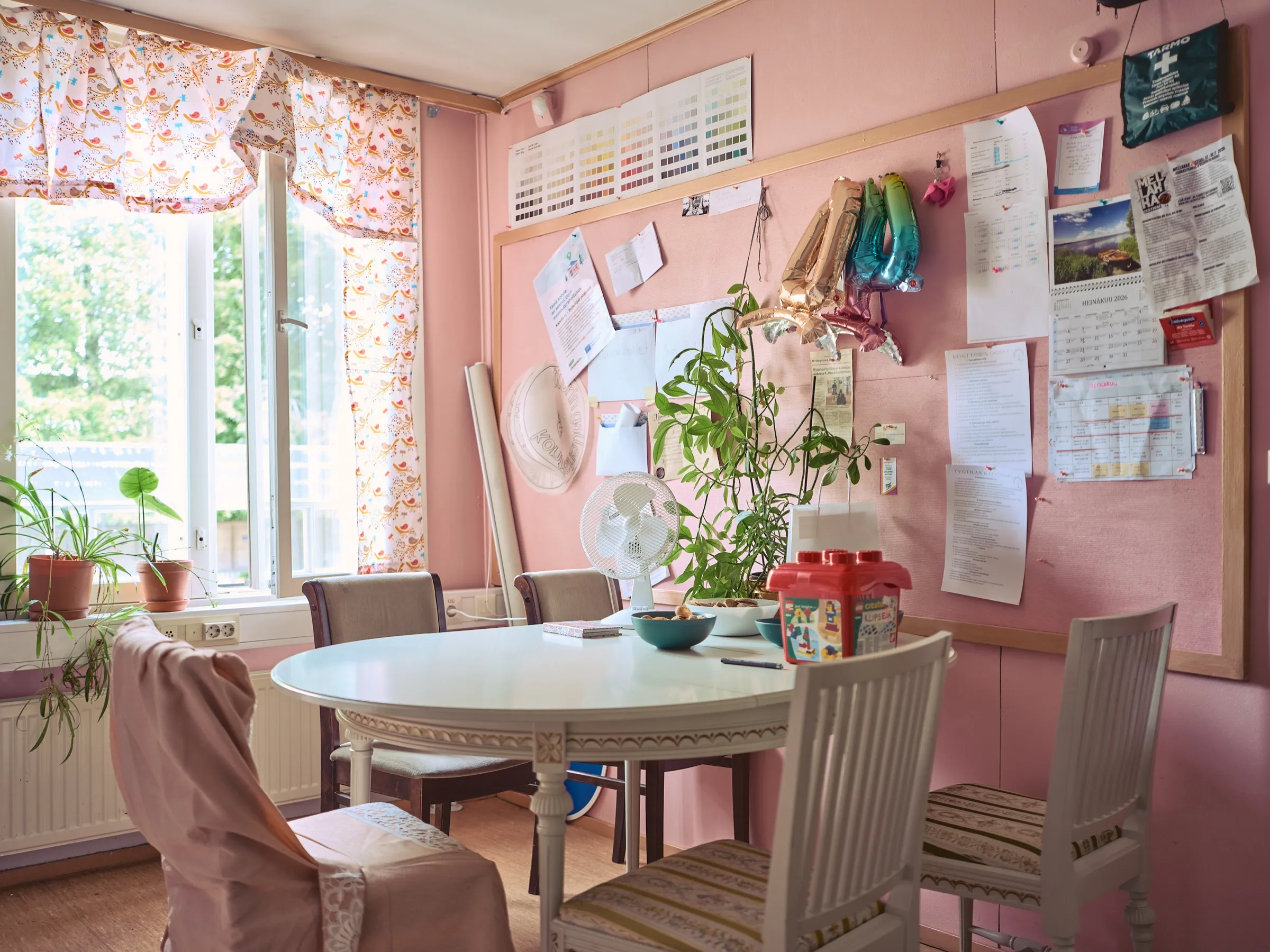 Rosa Besprechungsraum mit rundem weißem Tisch, großer Pinnwand und Zimmerpflanzen am Fenster