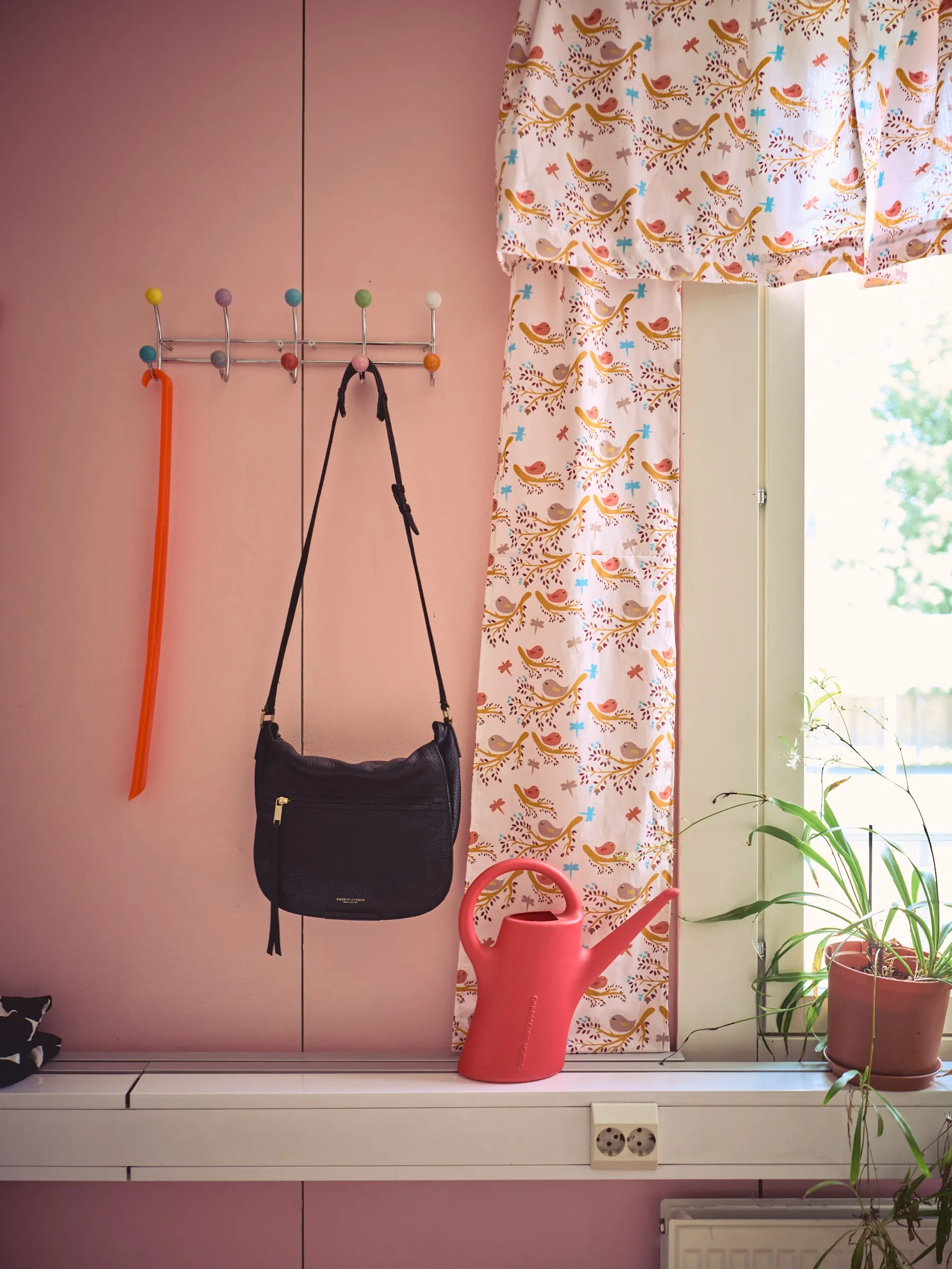 Rosa Wand mit bunter Hakenleiste, schwarzer Handtasche und roter Gießkanne am Fenster