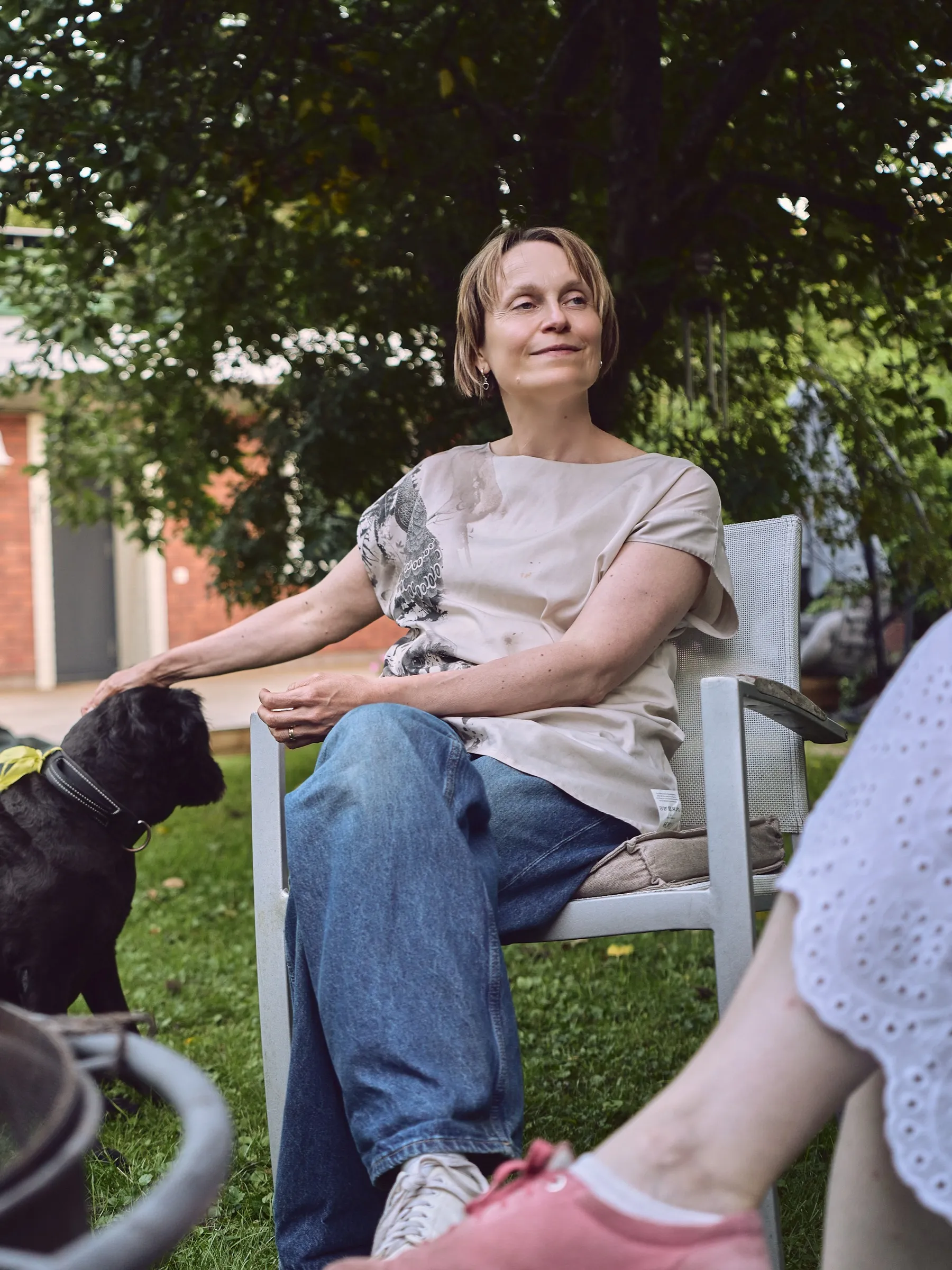 Frau sitzt entspannt auf einem Gartenstuhl unter Bäumen, neben ihr ein schwarzer Hund, Hochformat