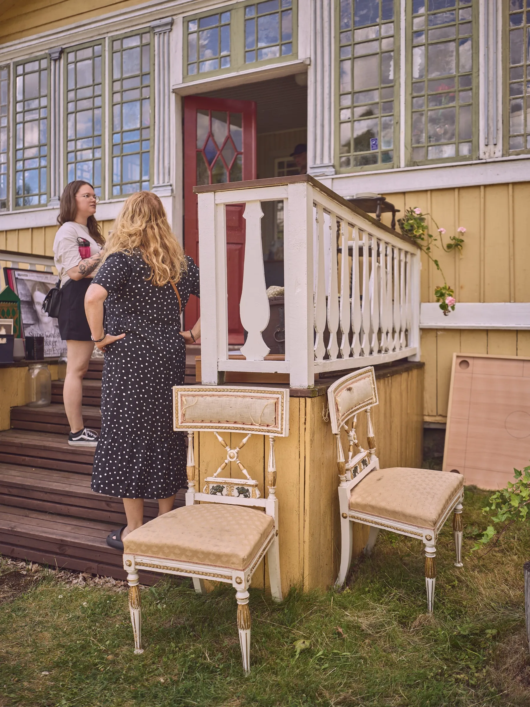 Zwei Frauen im Gespräch an der Treppe des gelben Hauses, davor zwei alte Polsterstühle, Hochformat