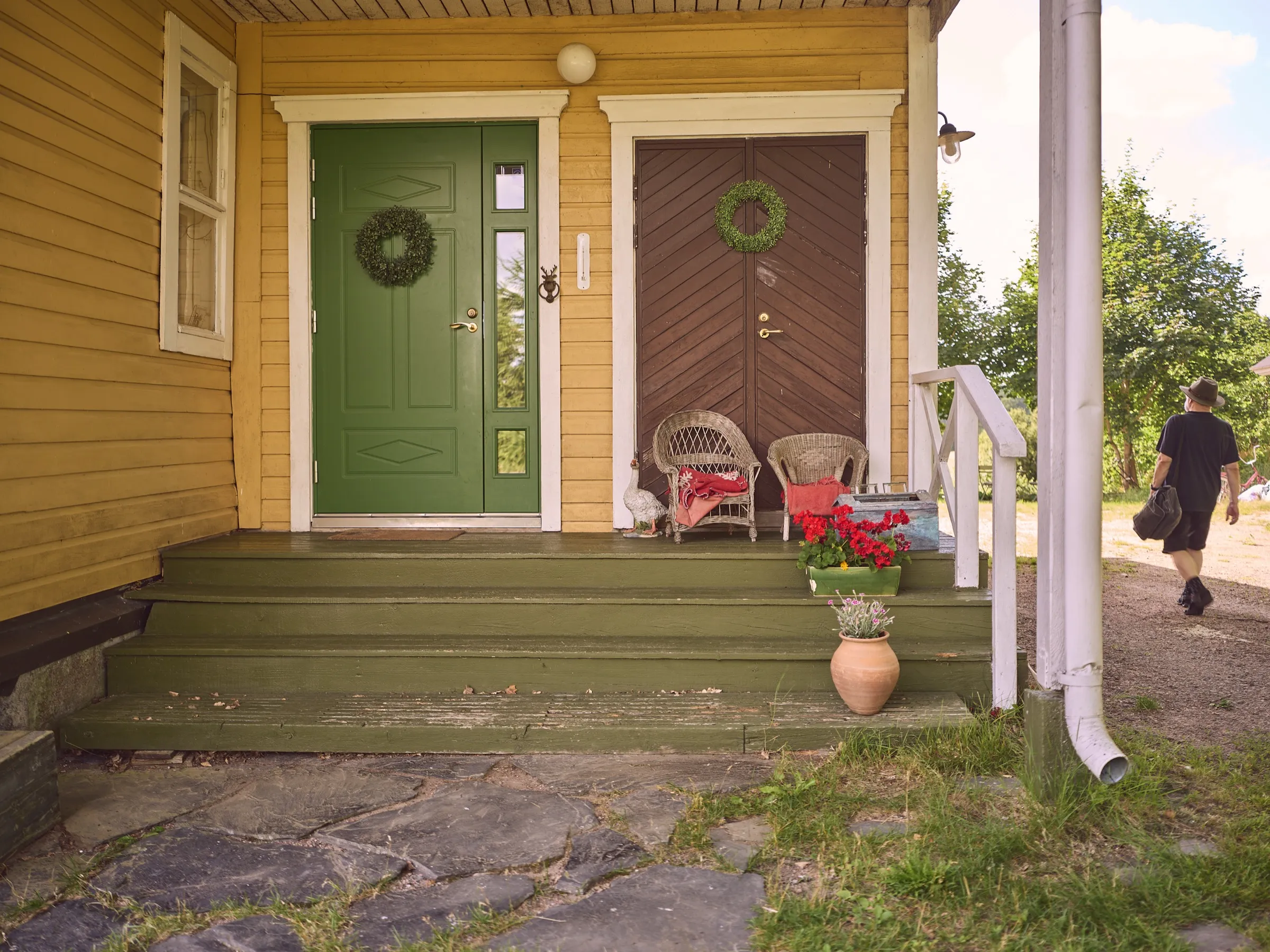 Veranda-Eingang des gelben Hauses mit grüner und brauner Tür, Kränzen und Korbstühlen