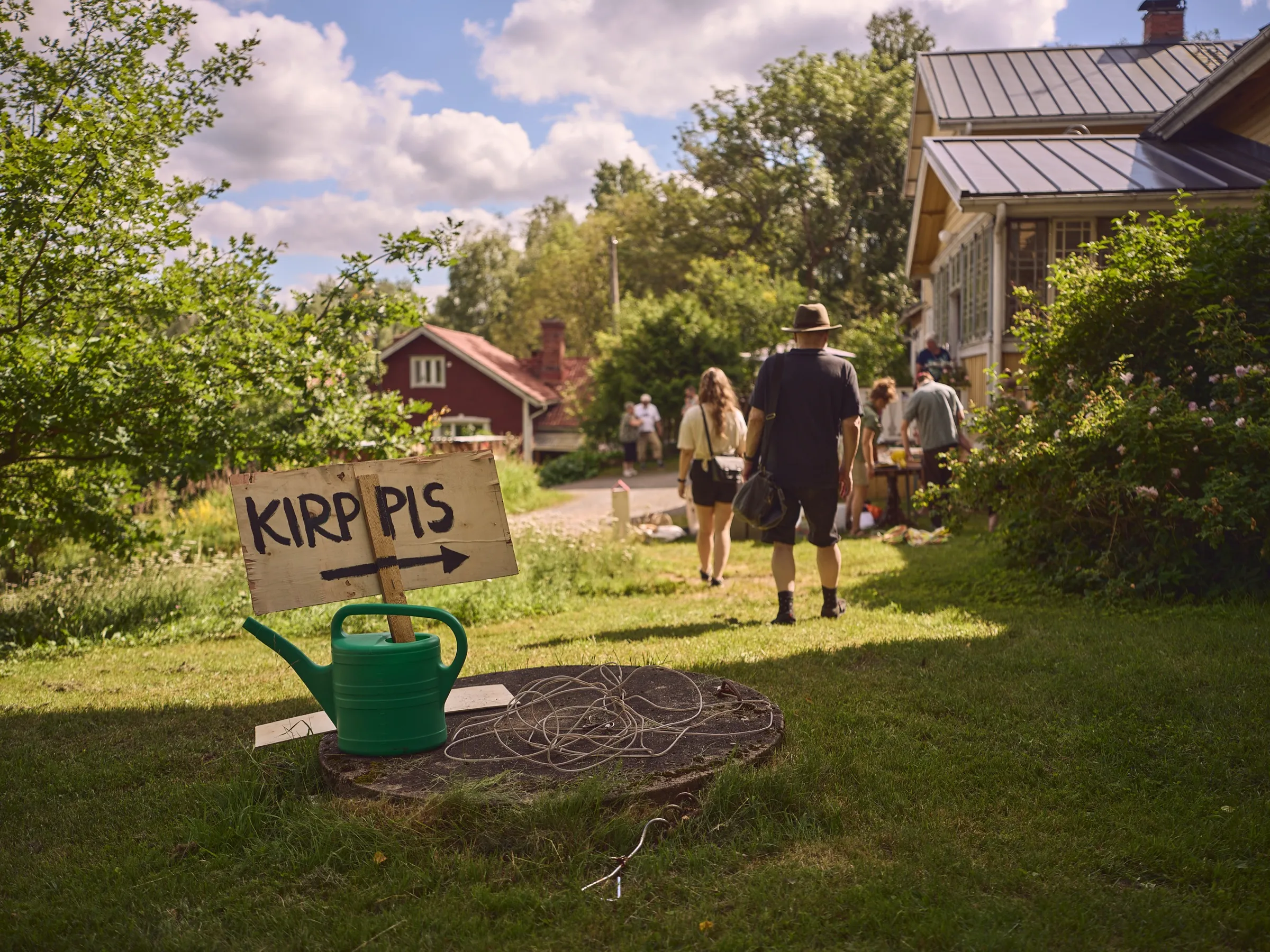 Handgemaltes KIRPPIS-Schild mit Pfeil auf einer grünen Gießkanne, dahinter gehen Menschen zum Hof