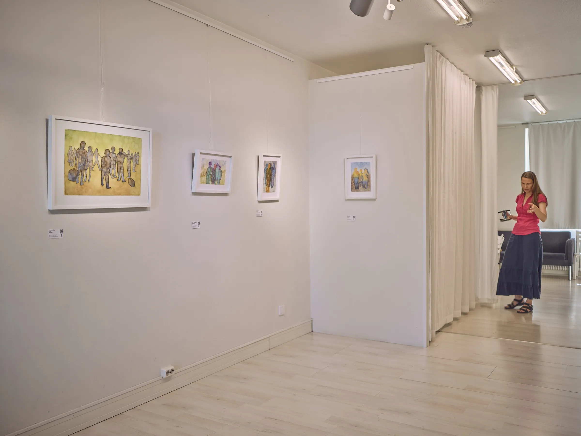 Weiße Galeriewand mit kleinen Aquarellen, im Hintergrund eine Frau am Vorhang