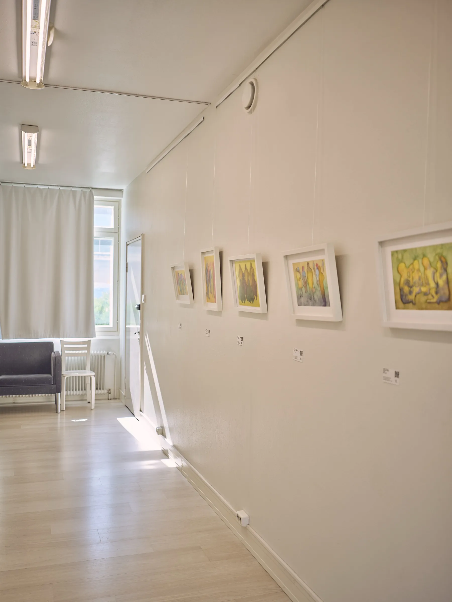 Lange helle Wand mit einer Reihe kleiner gerahmter Aquarelle, hinten ein blaues Sofa, Hochformat