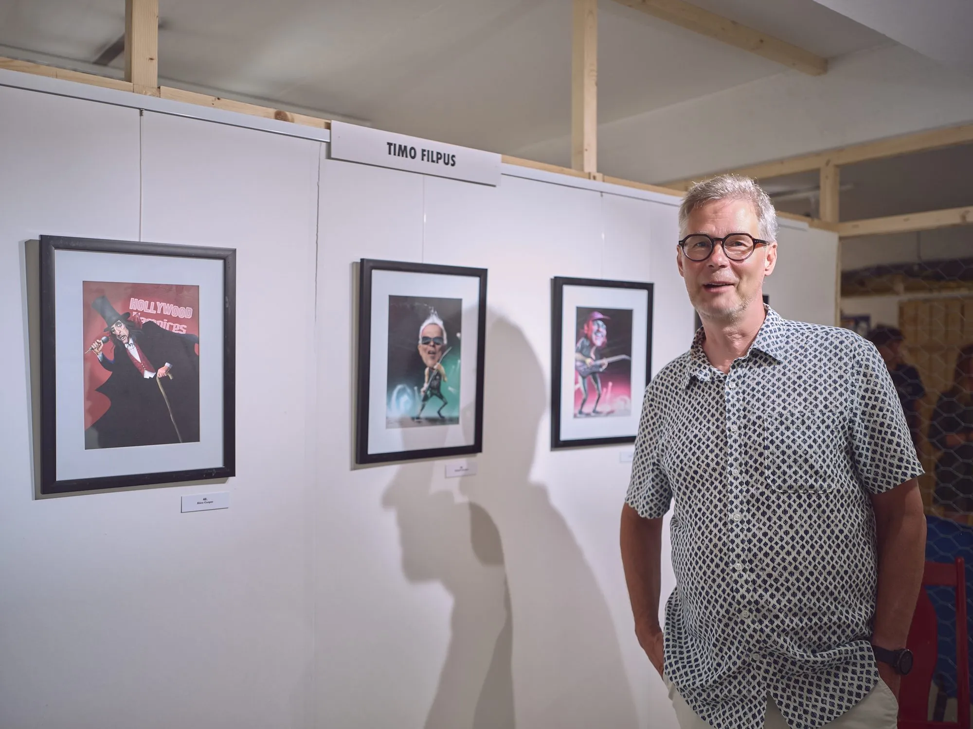Timo Filpus steht lächelnd an seiner Ausstellungswand mit gerahmten Musiker-Karikaturen