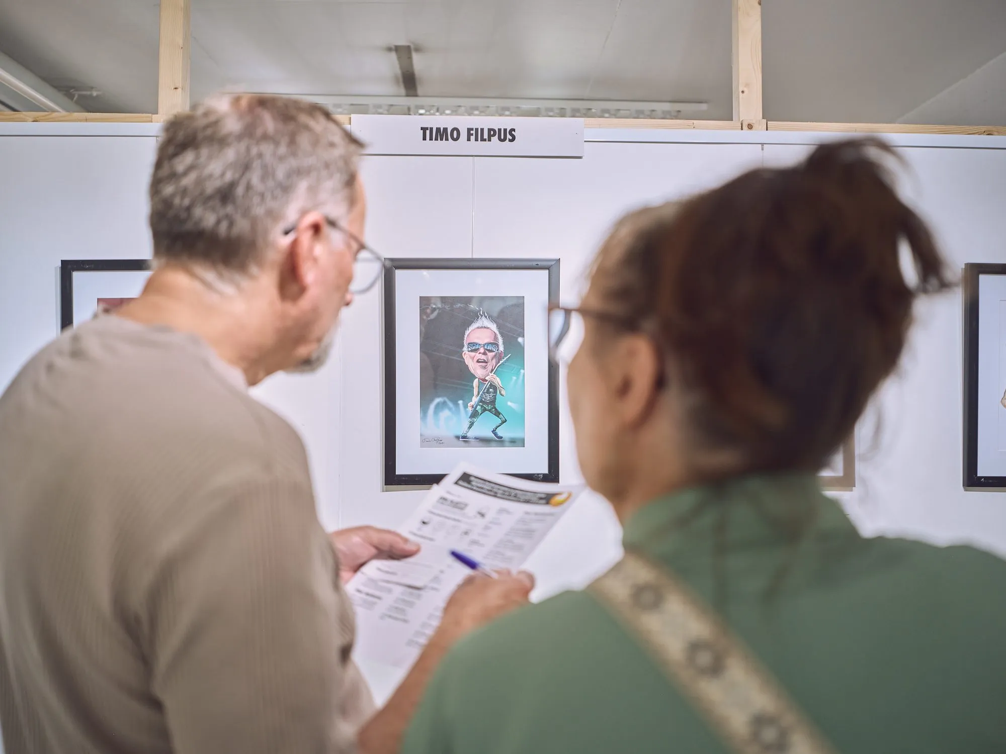 Zwei Besucher betrachten eine Musiker-Karikatur, einer hält die Werkliste in der Hand
