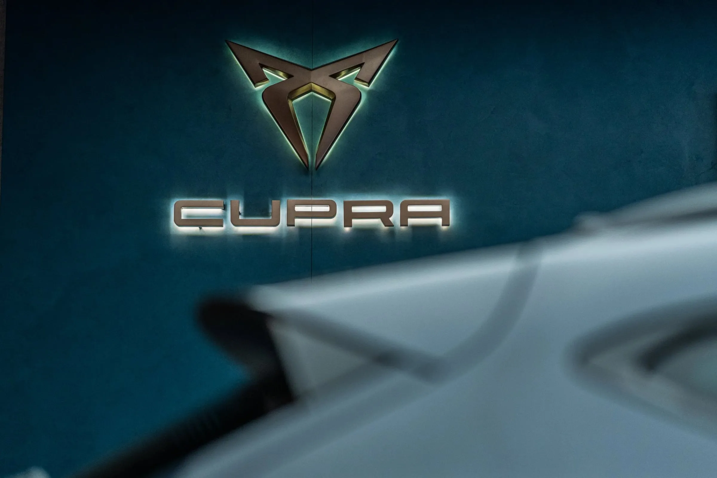 Beleuchtetes Cupra-Logo aus Metall auf petrolblauer Wand, davor unscharf eine helle Karosserie, dunkler Innenraum
