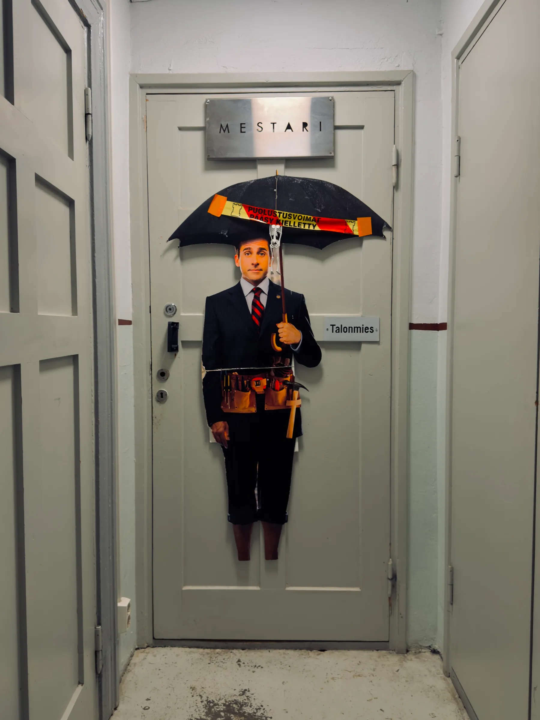 Hausmeistertür mit dem Schild Talonmies, davor ein Pappaufsteller mit aufgespanntem Schirm