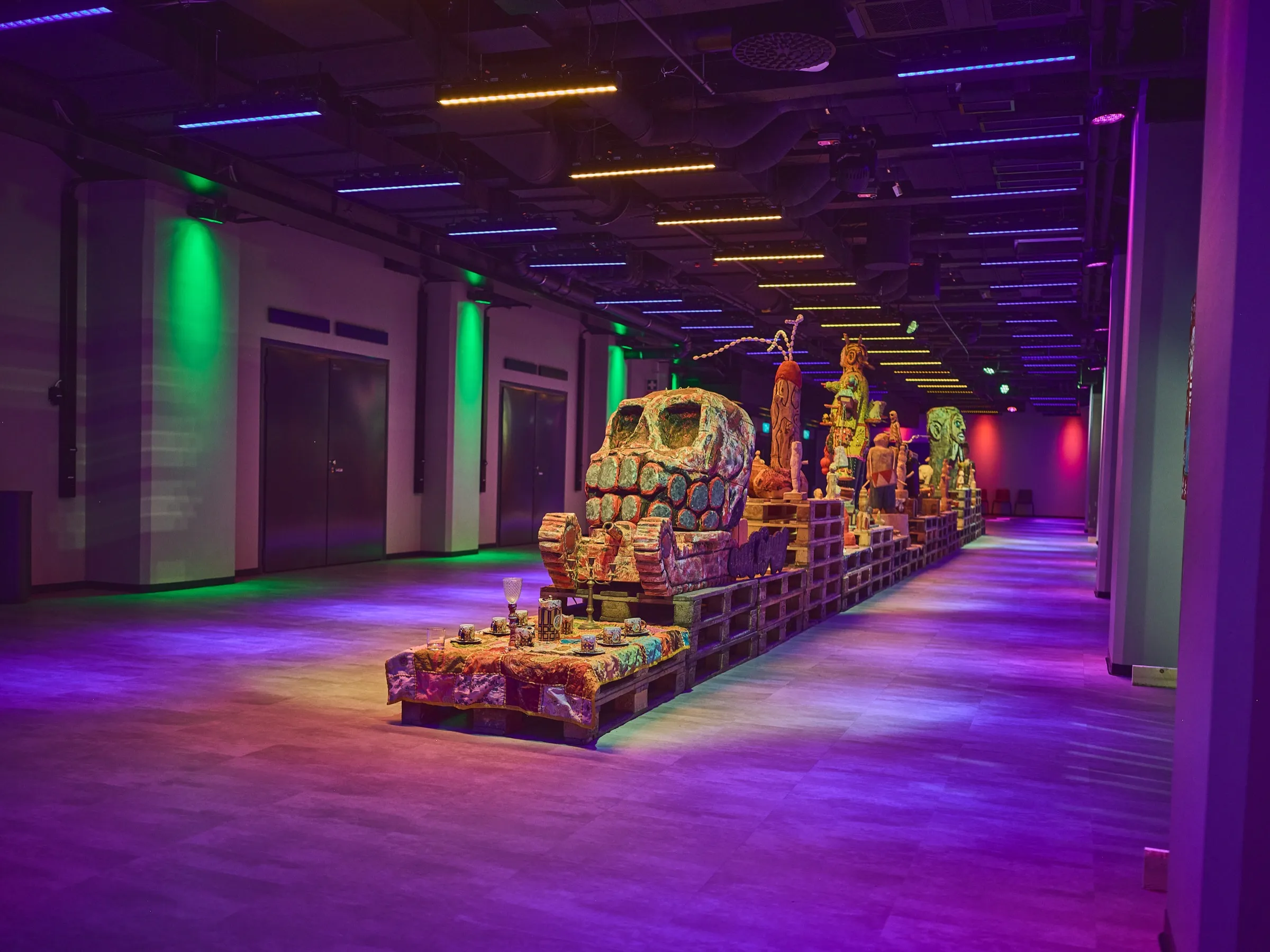 Weiter Blick durch den Saal, Holzfiguren auf Paletten entlang der Wand, blaues und violettes Licht