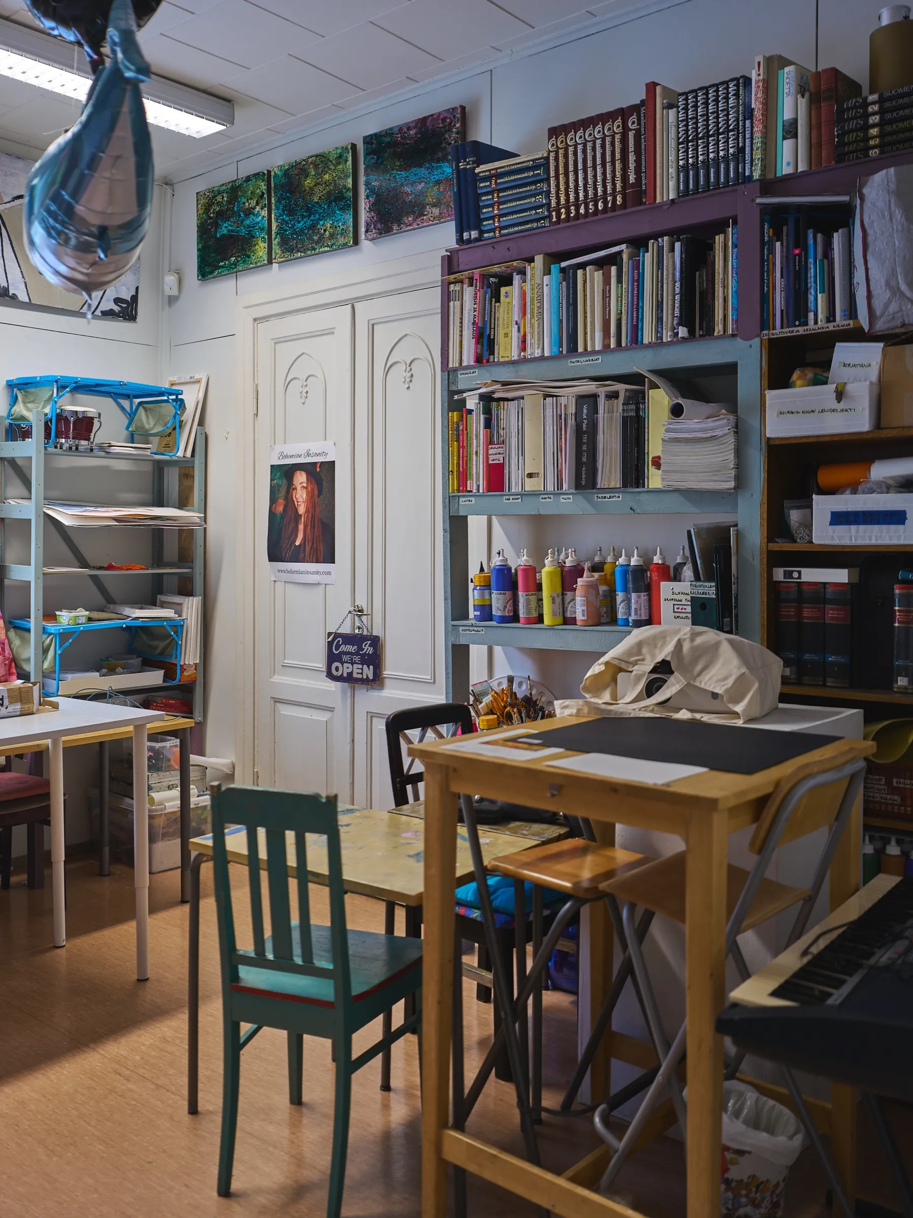 Werkraum mit Farbflaschen, Bücherregalen, Keyboard und Arbeitstischen unter drei kleinen Gemälden